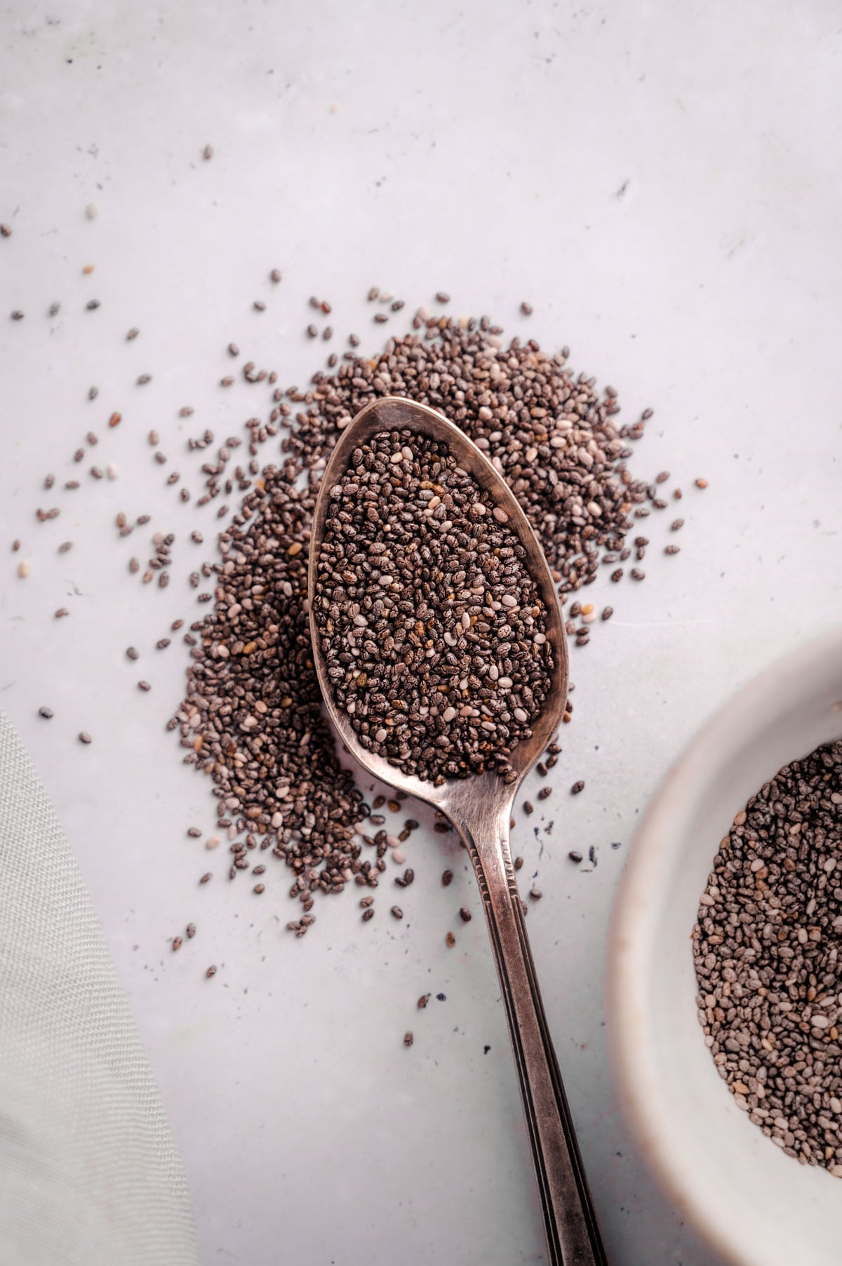 A spoon filled with chia seeds rests on a white surface, with additional scattered seeds around it. A bowl of chia seeds is partially visible in the corner.