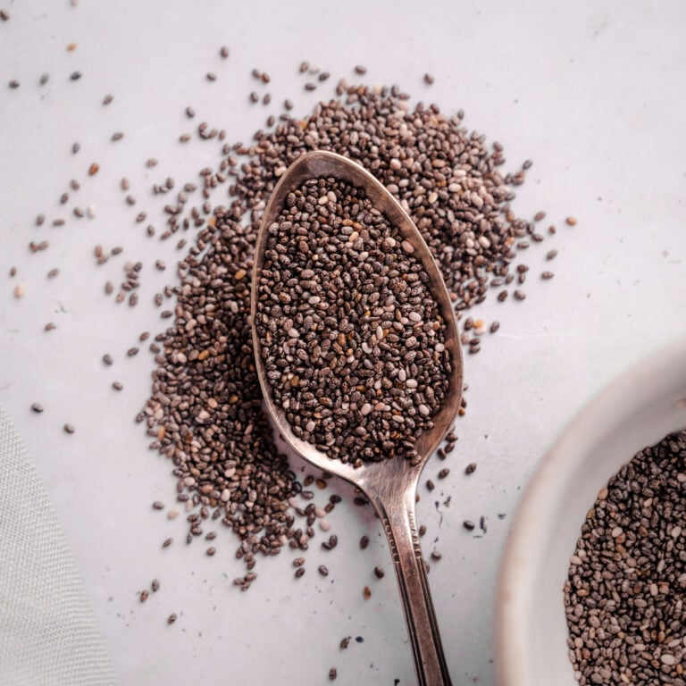 A wooden spoon filled with chia seeds rests on a white surface with additional chia seeds scattered around it. A white bowl partly filled with chia seeds is visible in the corner.
