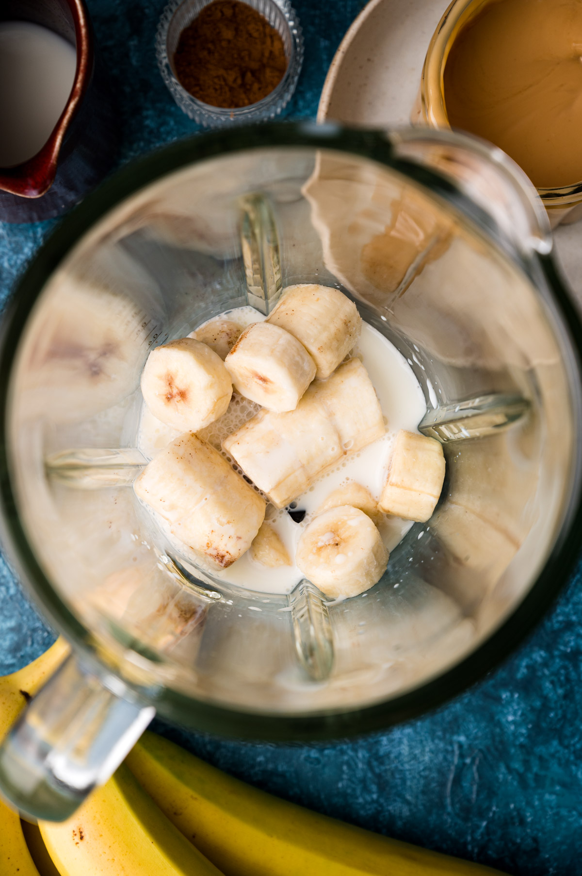 Close-up of a blender containing sliced bananas and milk, surrounded by ingredients such as cinnamon in a bowl, almond butter on a plate, and bananas on a blue surface.