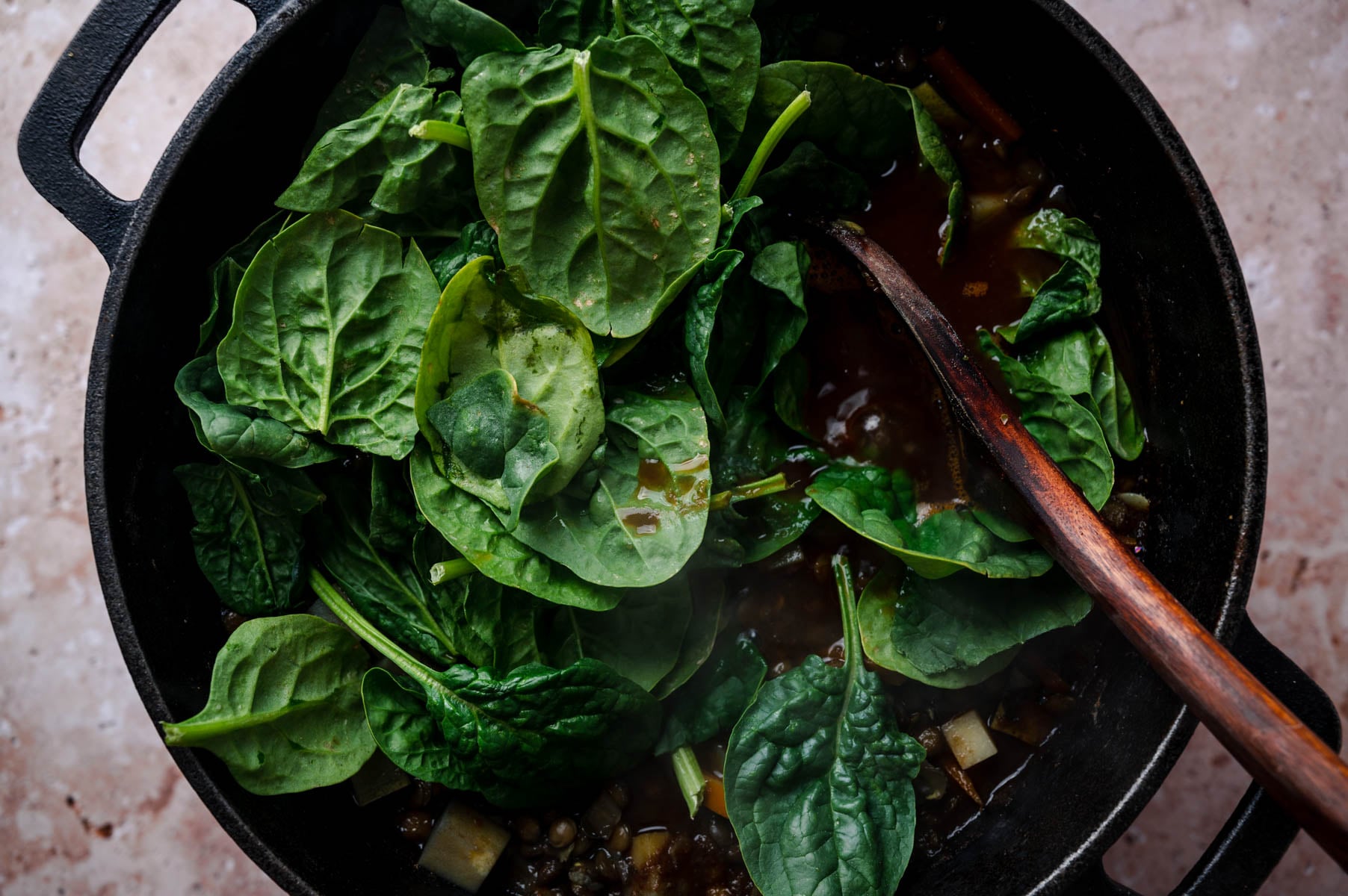 A large black pot filled with a leafy green vegetable, likely spinach, being cooked with a wooden spoon partially submerged in the mixture.