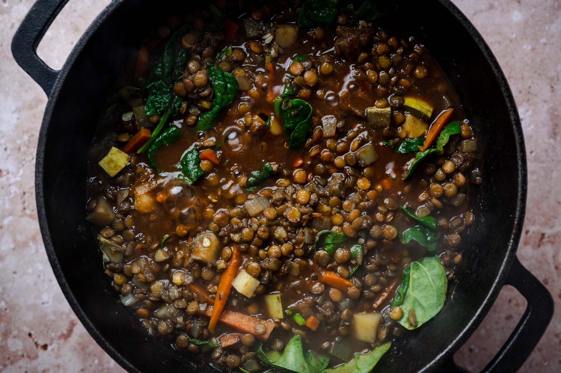 A black pot filled with a mixed vegetable and lentil stew, featuring visible spinach leaves, carrots, and diced potatoes.