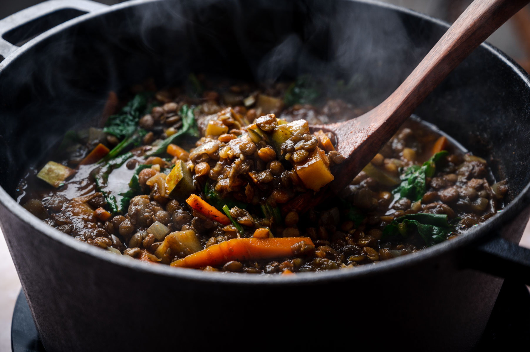 Close-up of a steaming pot of vegetable soup, with a wooden spoon stirring ingredients like lentils, carrots, greens, and other vegetables.