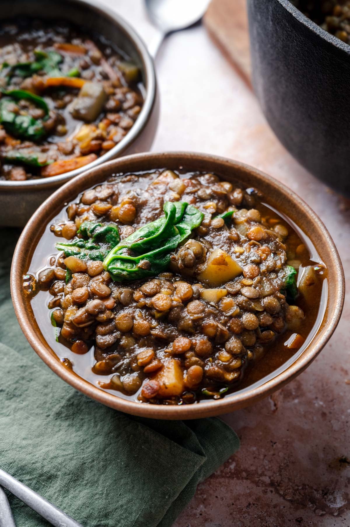 A close-up of a hearty lentil soup in a bowl, garnished with fresh greens. Another bowl of soup and a silver spoon are partially visible in the background.