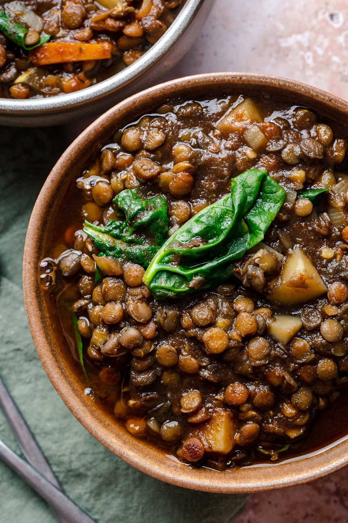 A bowl of lentil soup with spinach and chopped vegetables, placed on a tablecloth with a spoon nearby.