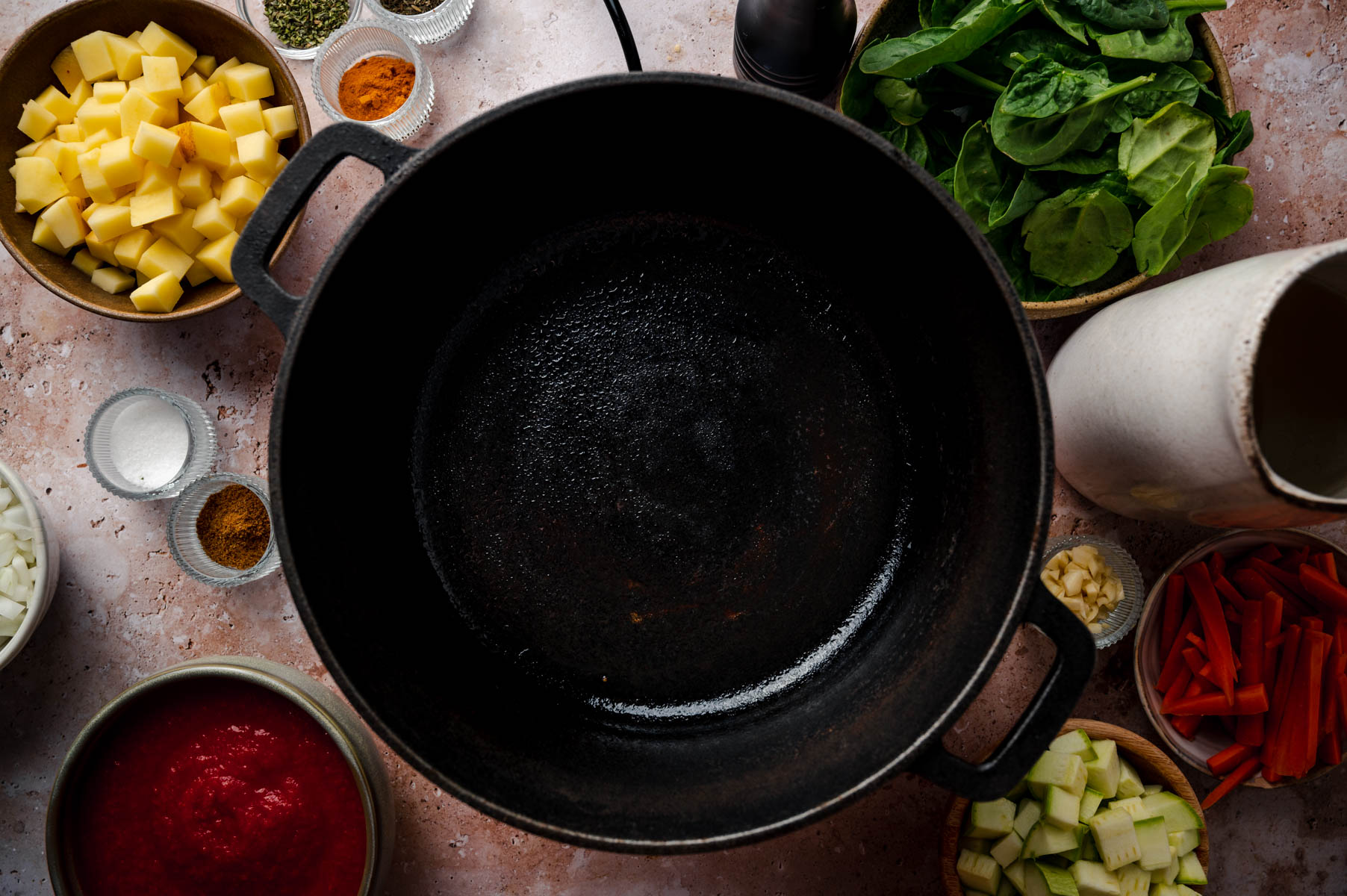 A large black pot centered on a countertop, surrounded by bowls containing diced vegetables, spices, and a container with liquid.