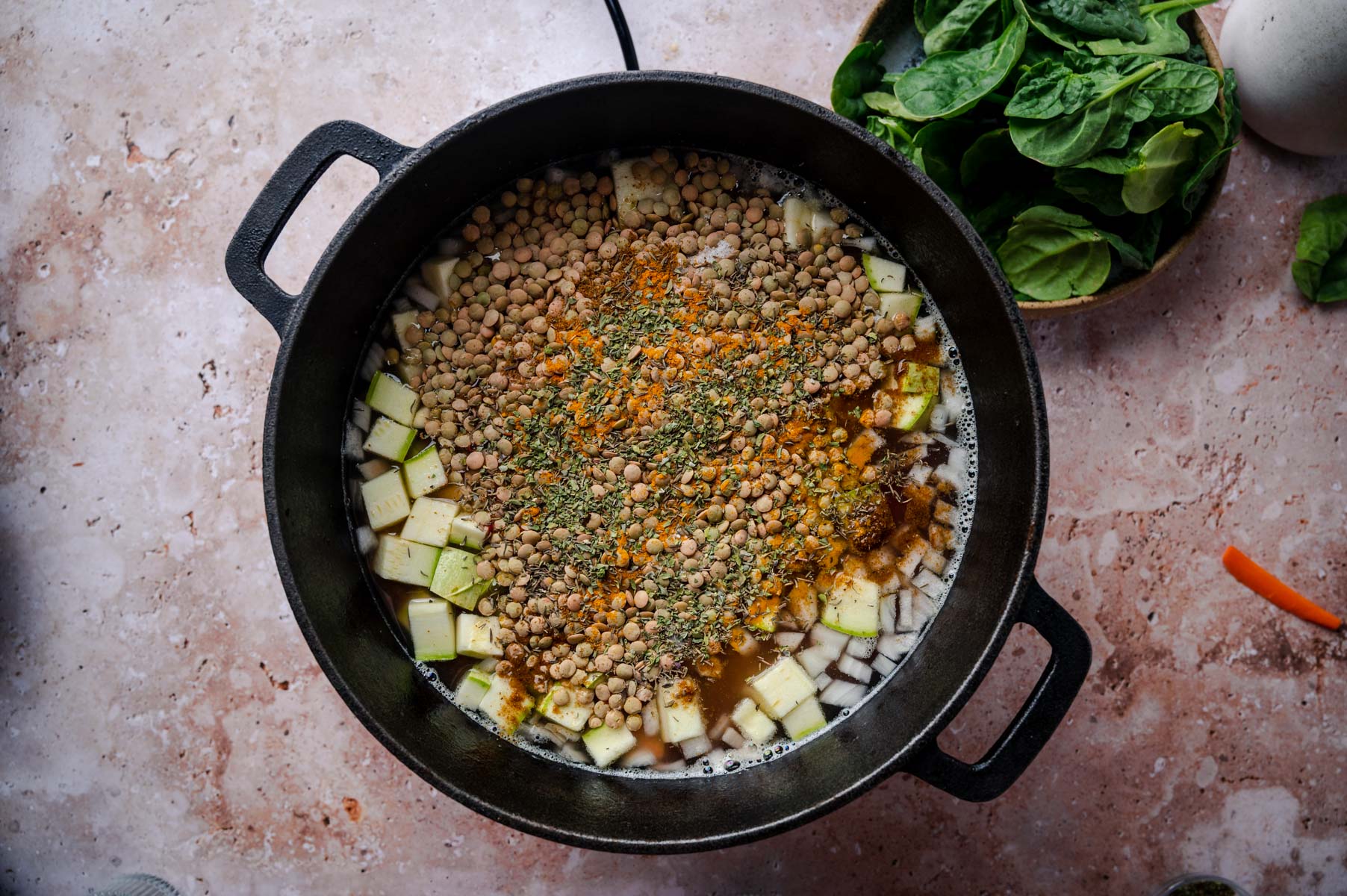 A black Dutch oven on a pinkish countertop contains a soup mixture of chopped vegetables, lentils, and spices. Fresh spinach leaves are seen in a bowl next to the pot.