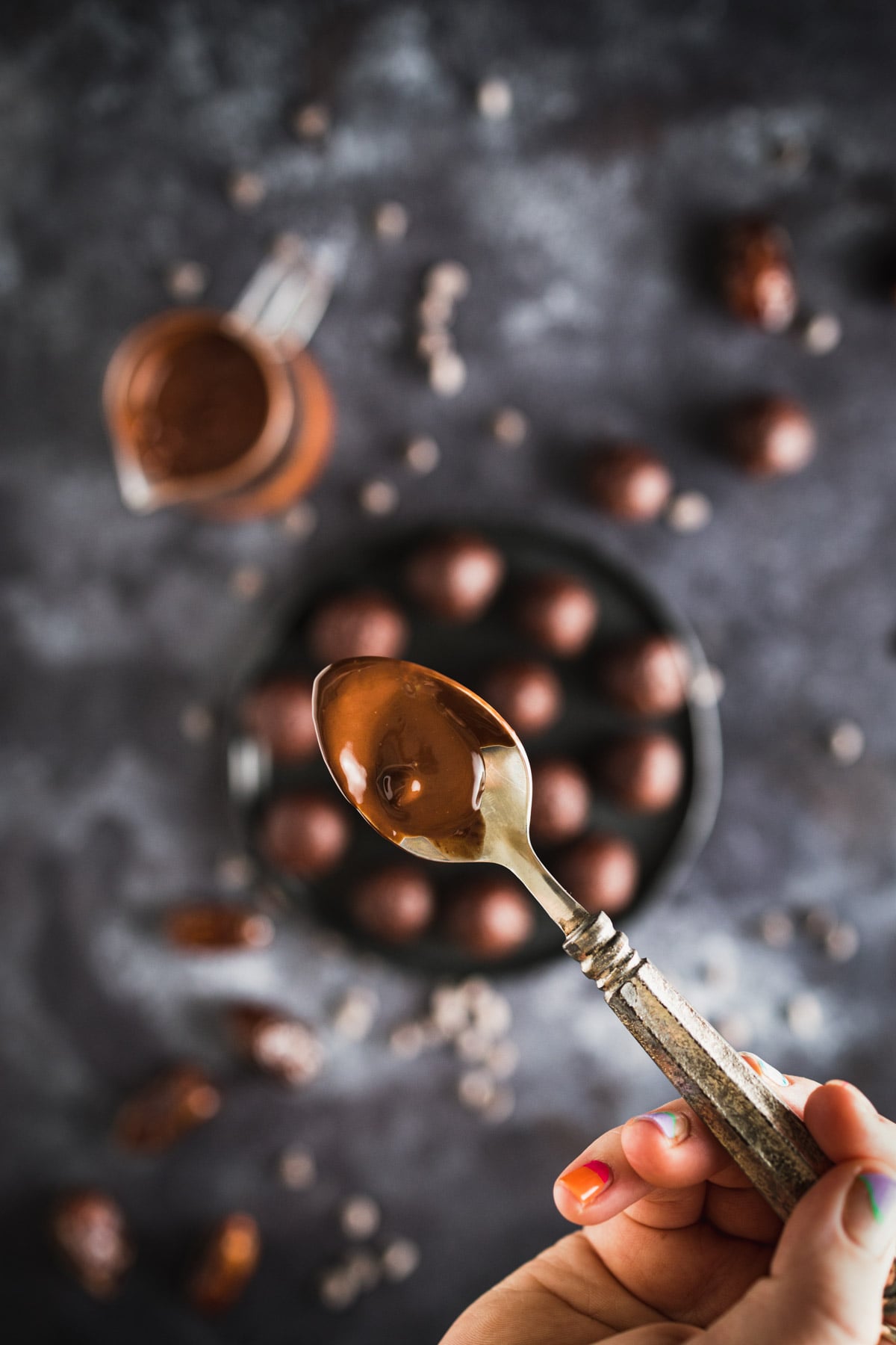 Close-up of a hand holding a spoon with melted chocolate. In the blurred background, there are round chocolate truffles on a black plate, small scattered chocolate pieces, and a small jar of melted chocolate.