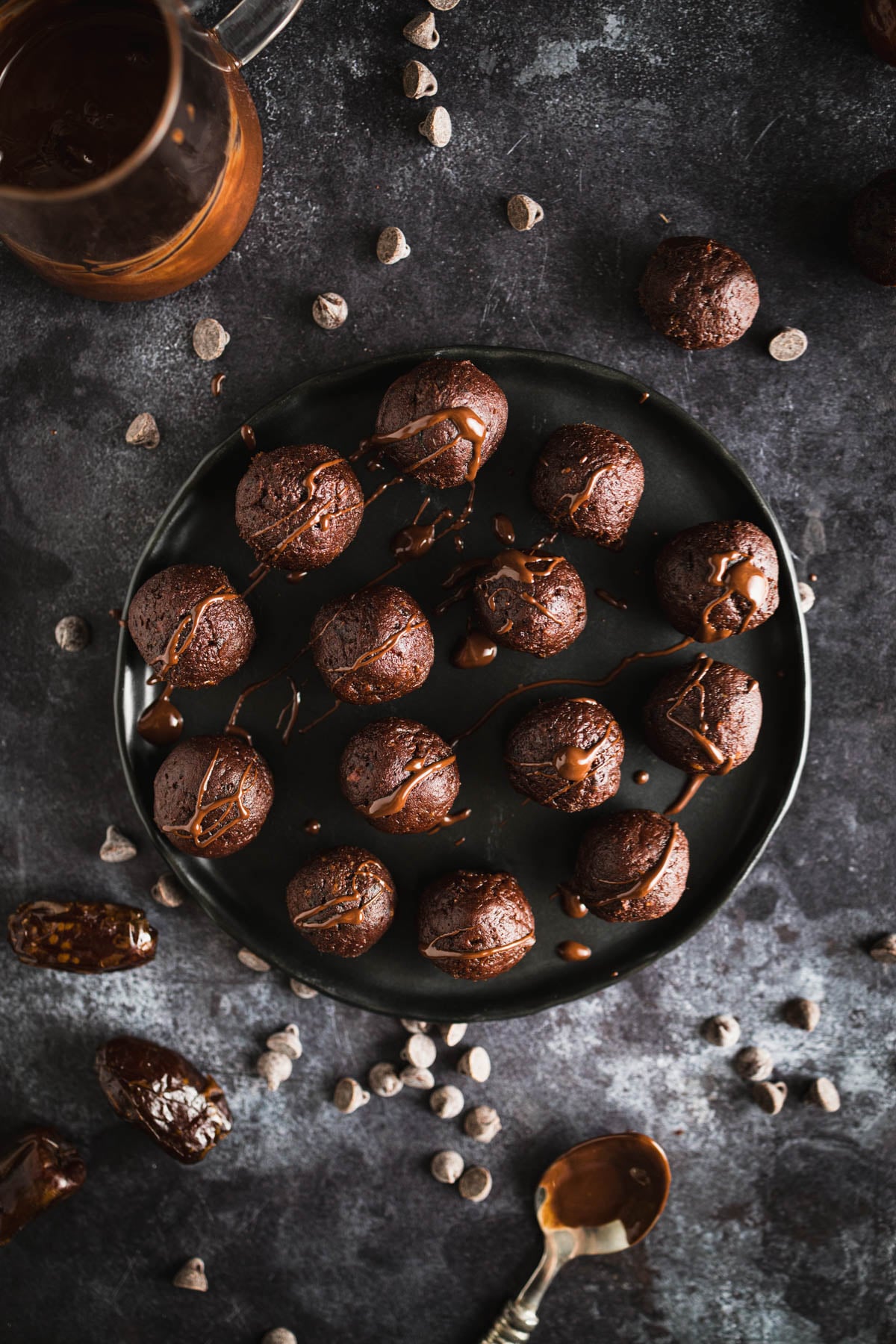 A black plate with chocolate date balls drizzled with chocolate sauce, surrounded by scattered chocolate chips and dates on a dark textured surface. A spoon and a glass jug with a melted chocolate are nearby.