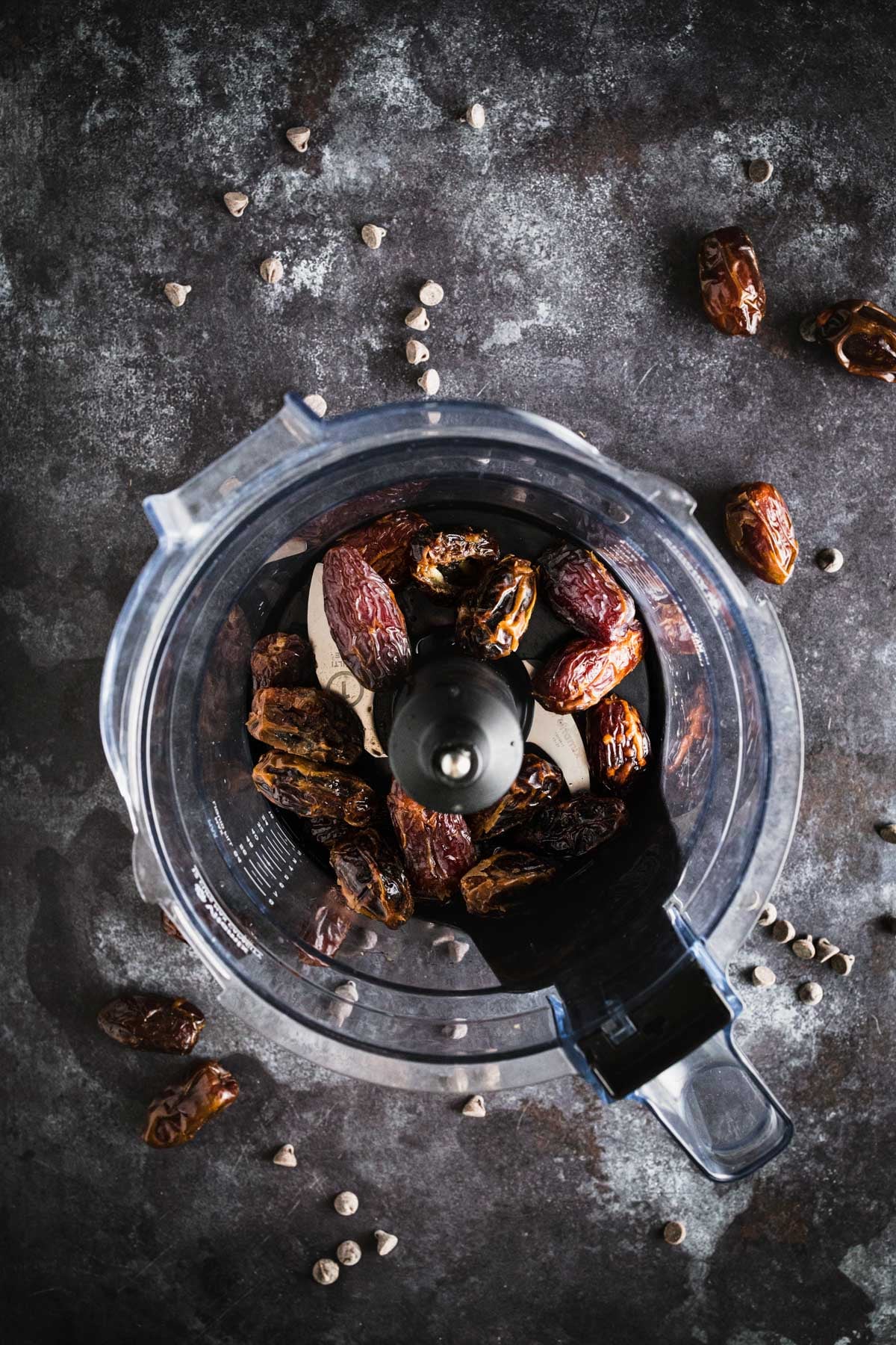 A food processor bowl containing pitted dates, set on a dark textured surface with a few scattered dates and chocolate chips around it.