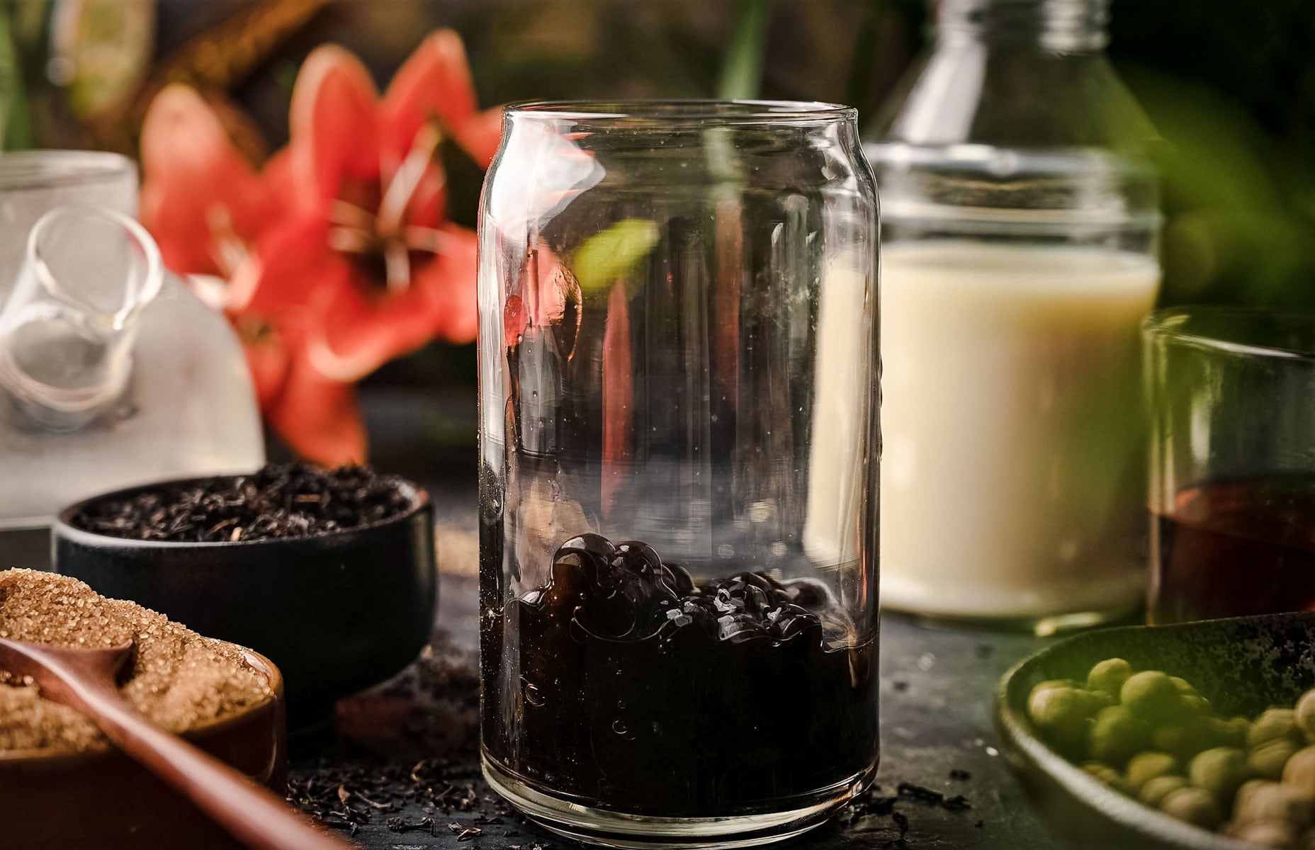 A transparent glass jar containing dark tapioca pearls is surrounded by various ingredients, including brown sugar, tea leaves, milk, and a red flower in the background.