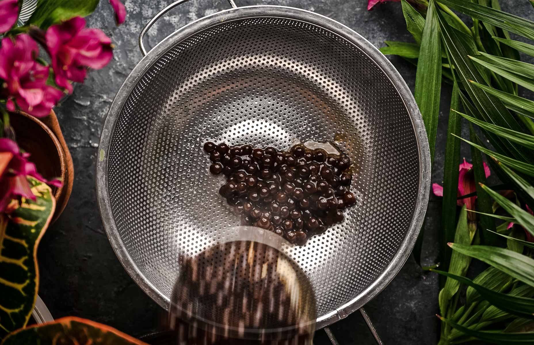 A metal colander containing a small cluster of boba on a gray surface, surrounded by pink flowers and green leaves.