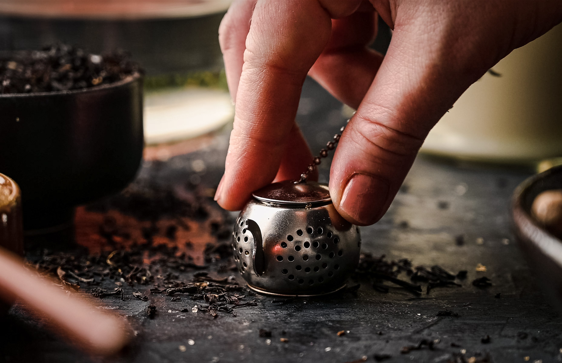 A hand is holding a small, round tea infuser on a dark surface with loose tea leaves scattered around. A bowl filled with tea leaves is in the background.