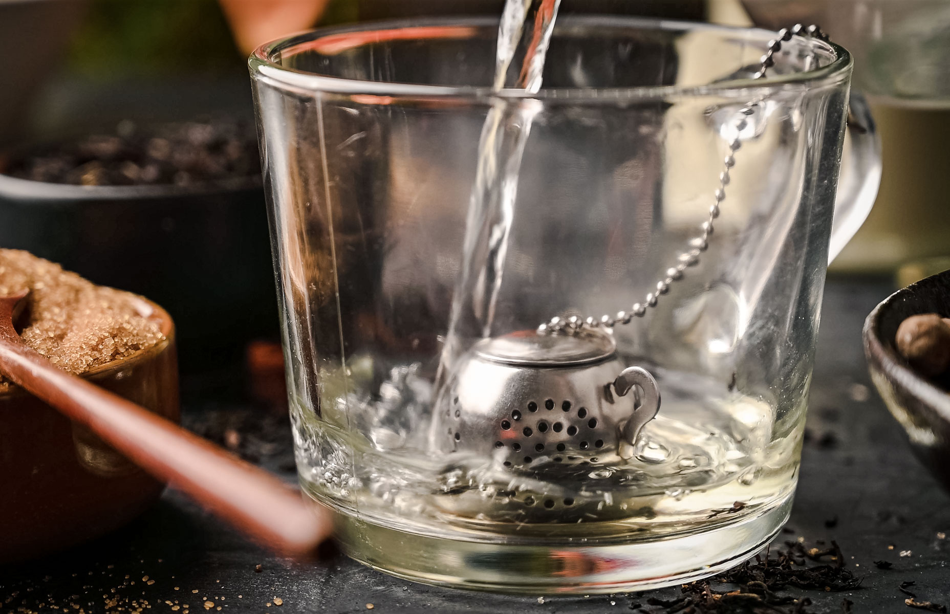Water being poured into a glass cup containing a teapot-shaped tea infuser on a chain. Tea ingredients surround the cup.