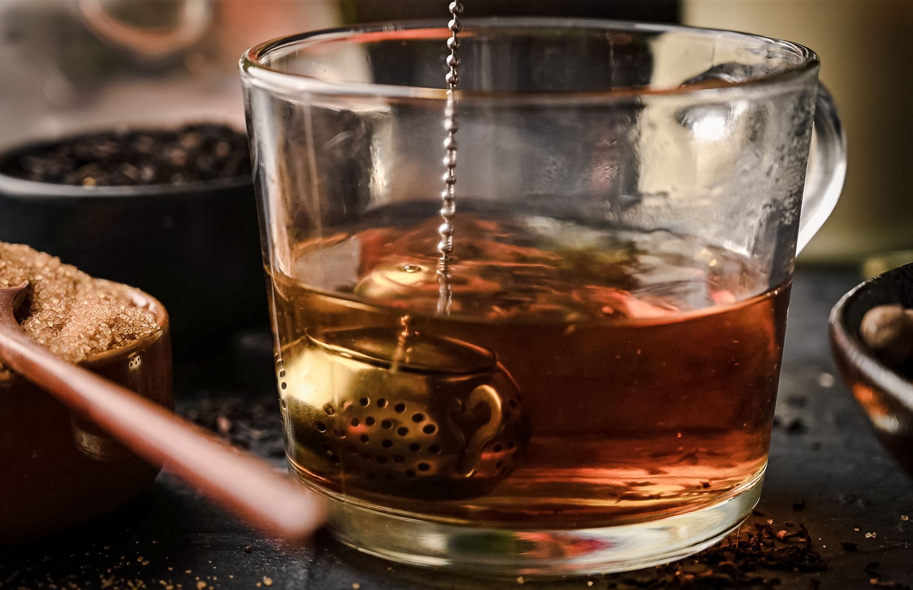 A close-up of a glass mug of tea with a metal tea infuser submerged in the liquid. Surrounding the mug are various containers of loose leaf tea and a spoon with honey.
