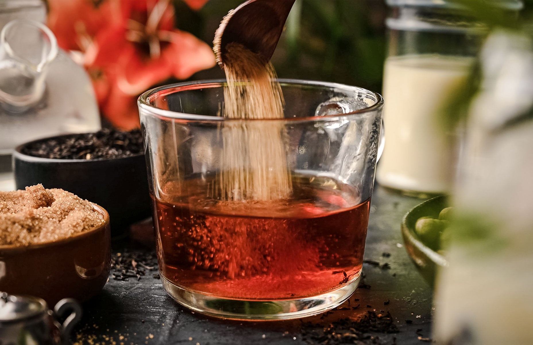 A wooden spoon pours sugar into a glass of dark brown liquid on a table with bowls of ingredients and a drink pitcher in the background.