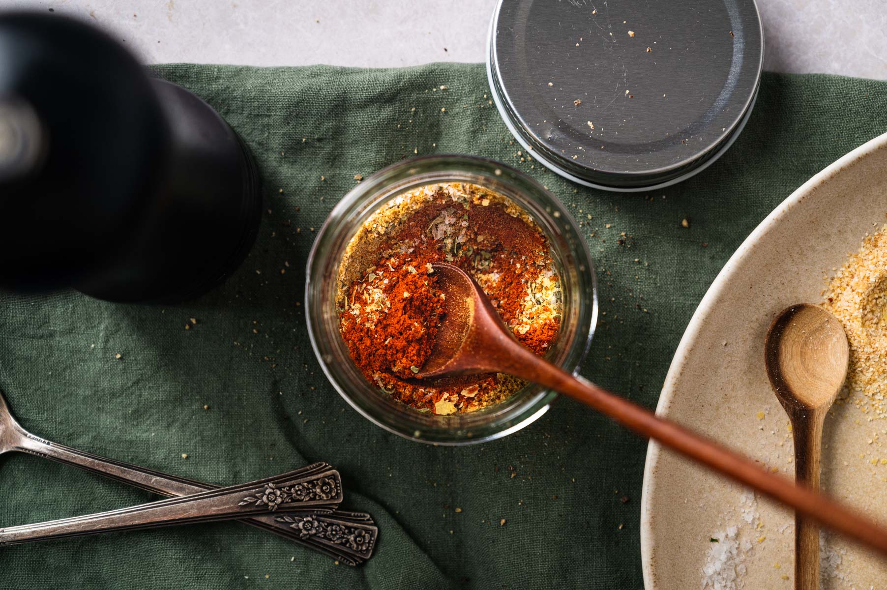 A wooden spoon in a jar filled with mixed spices on a green cloth; nearby are a plate with spoons and a pepper grinder.