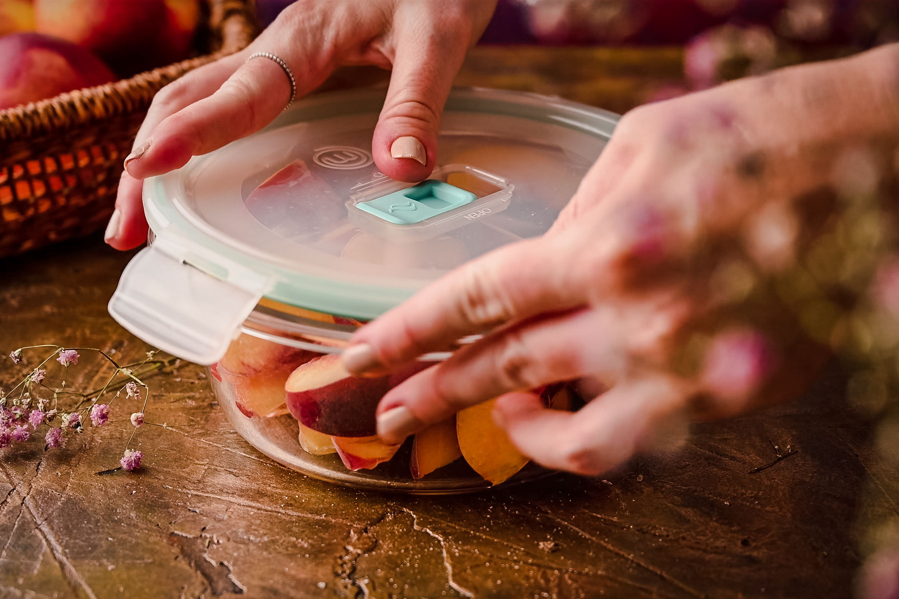 Hands sealing a glass container filled with sliced peaches using a lid. A basket with additional peaches is visible in the background on a wooden surface.