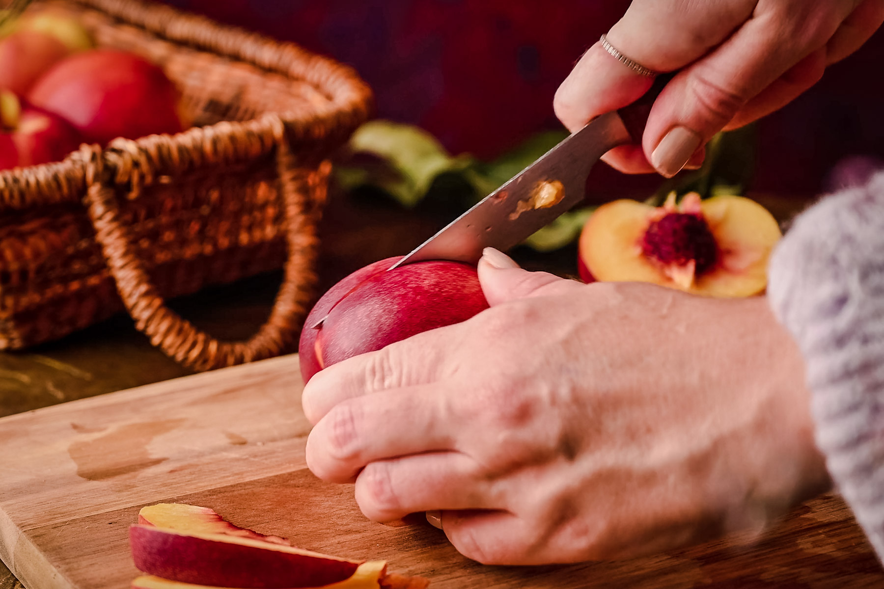 Hands are cutting a ripe peach with a knife on a wooden cutting board. A wicker basket with more peaches is in the background.
