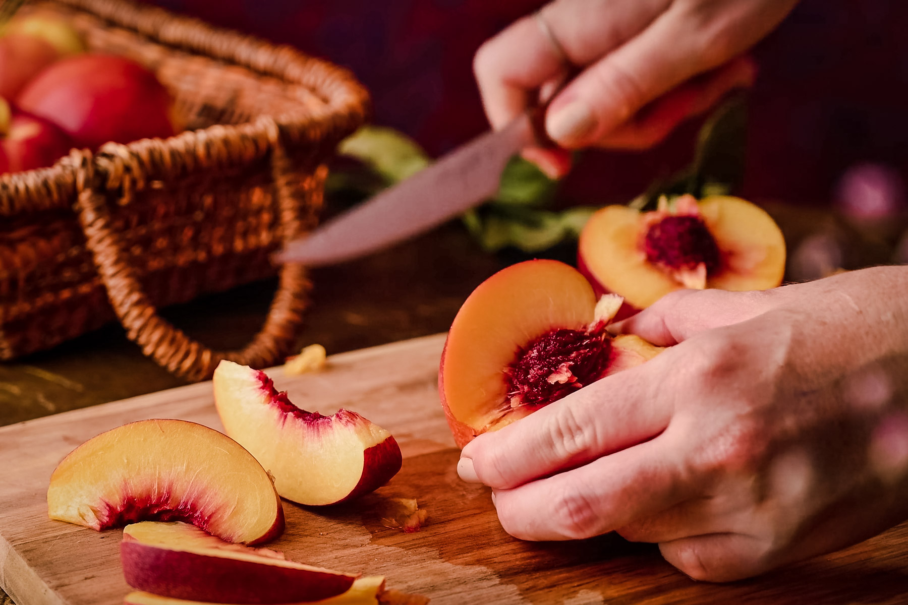 A person slices ripe peaches on a wooden cutting board. A woven basket containing more peaches is in the background.