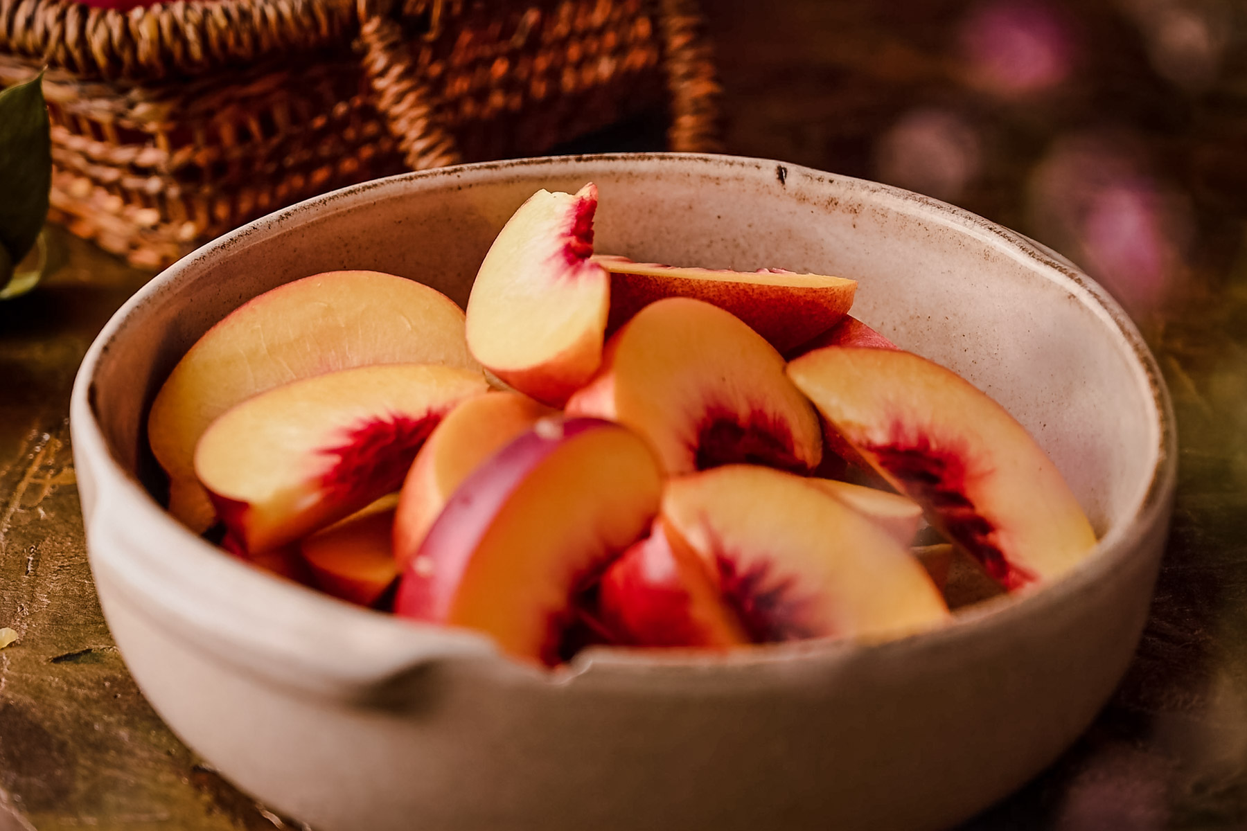 A ceramic bowl filled with sliced peaches is placed on a rustic surface with a wicker basket in the background.