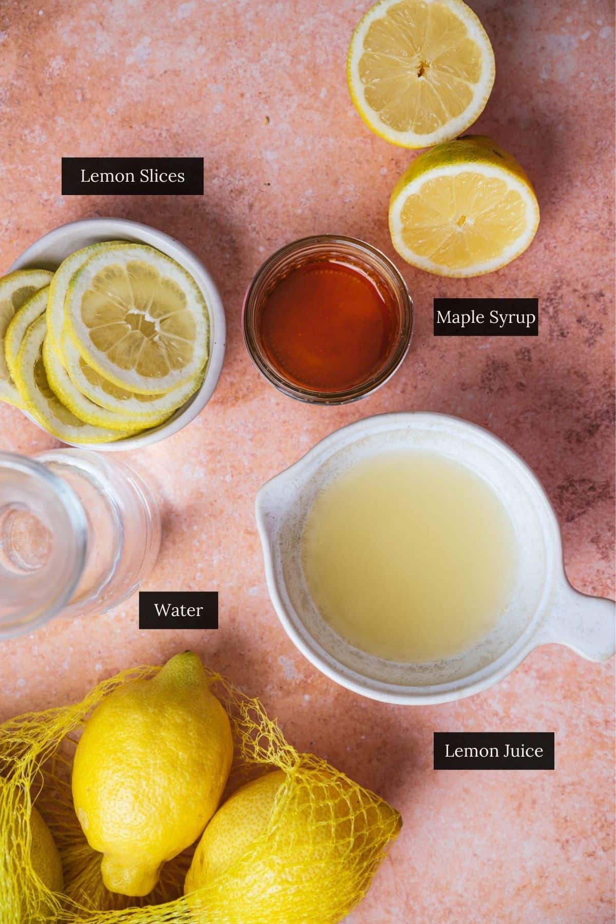 Ingredients displayed on a table: lemon slices in a bowl, halved lemons, a jar of maple syrup, a jar of water, lemon juice in a container, and a net containing whole lemons.