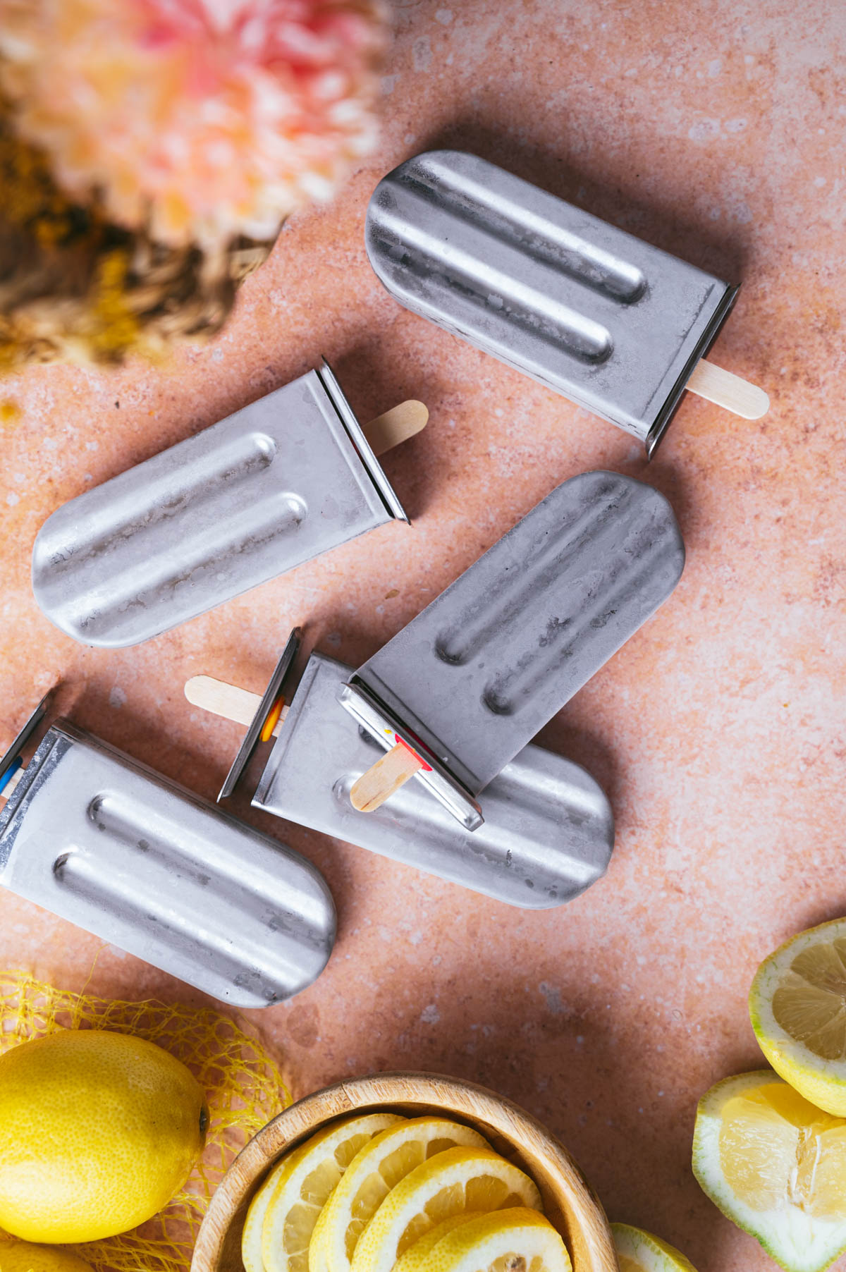 Metal popsicle molds laid on a pink surface with lemons and lemon slices nearby.