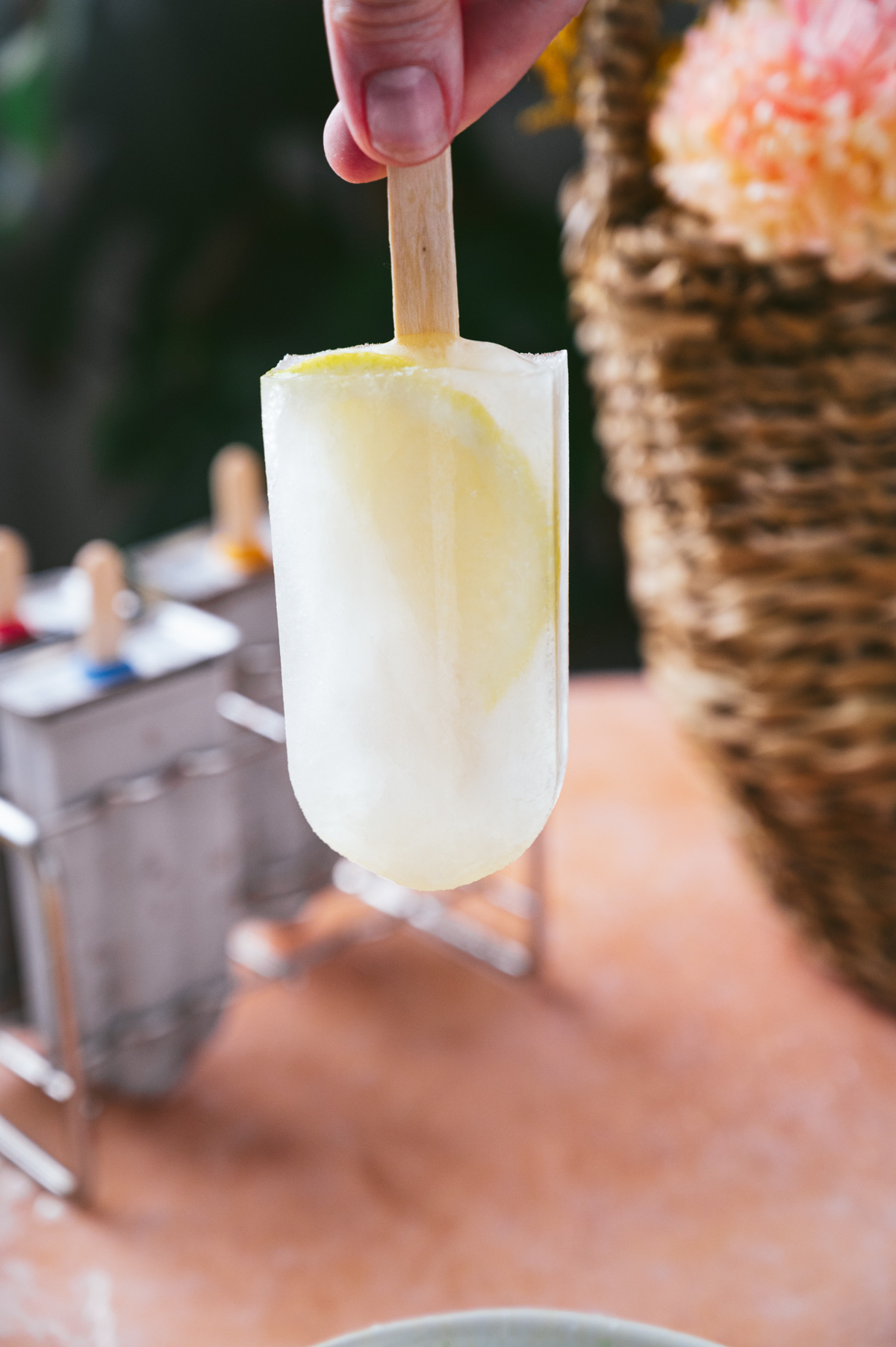 A hand holding a lemon slice popsicle against a blurred background with other popsicles in a metal holder and a wicker basket.