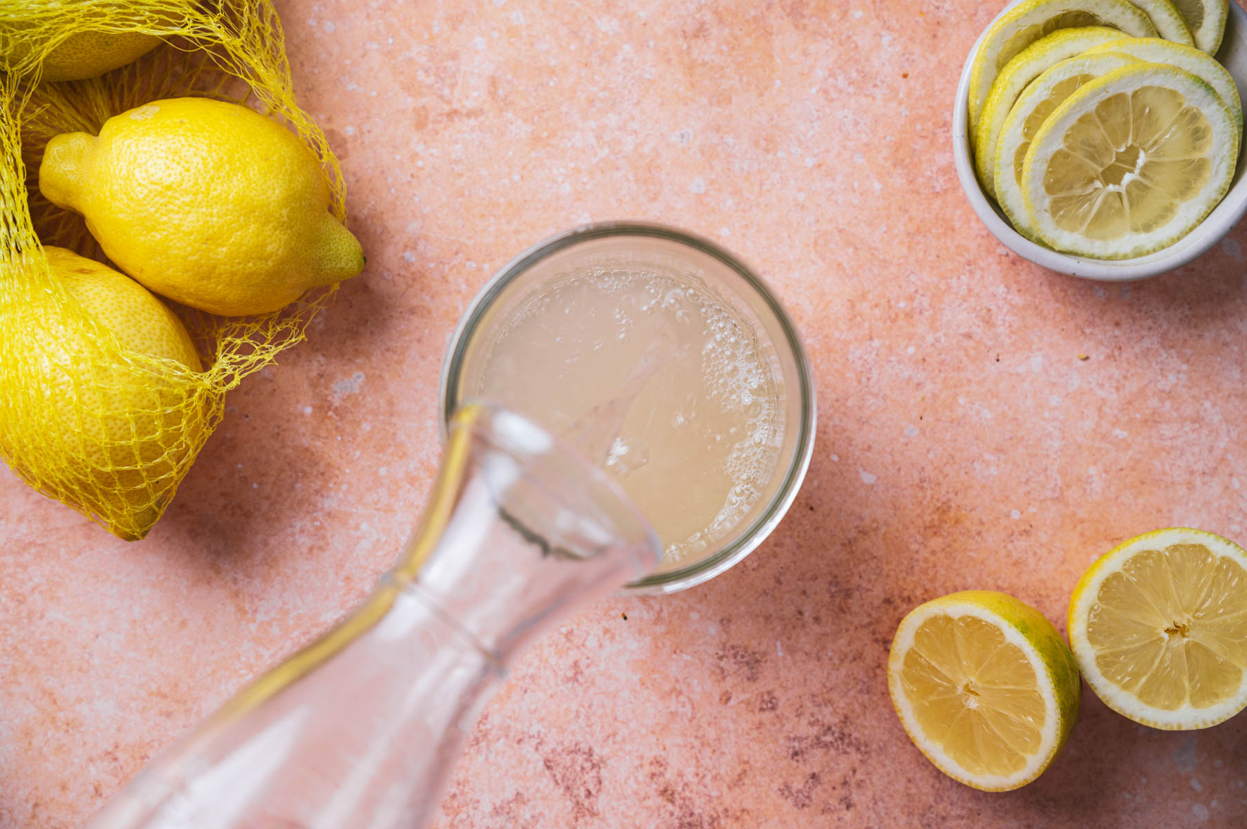 A glass is being filled with lemon juice from a clear bottle, surrounded by a bag of whole lemons, a bowl of lemon slices, and halved lemons on a textured surface.