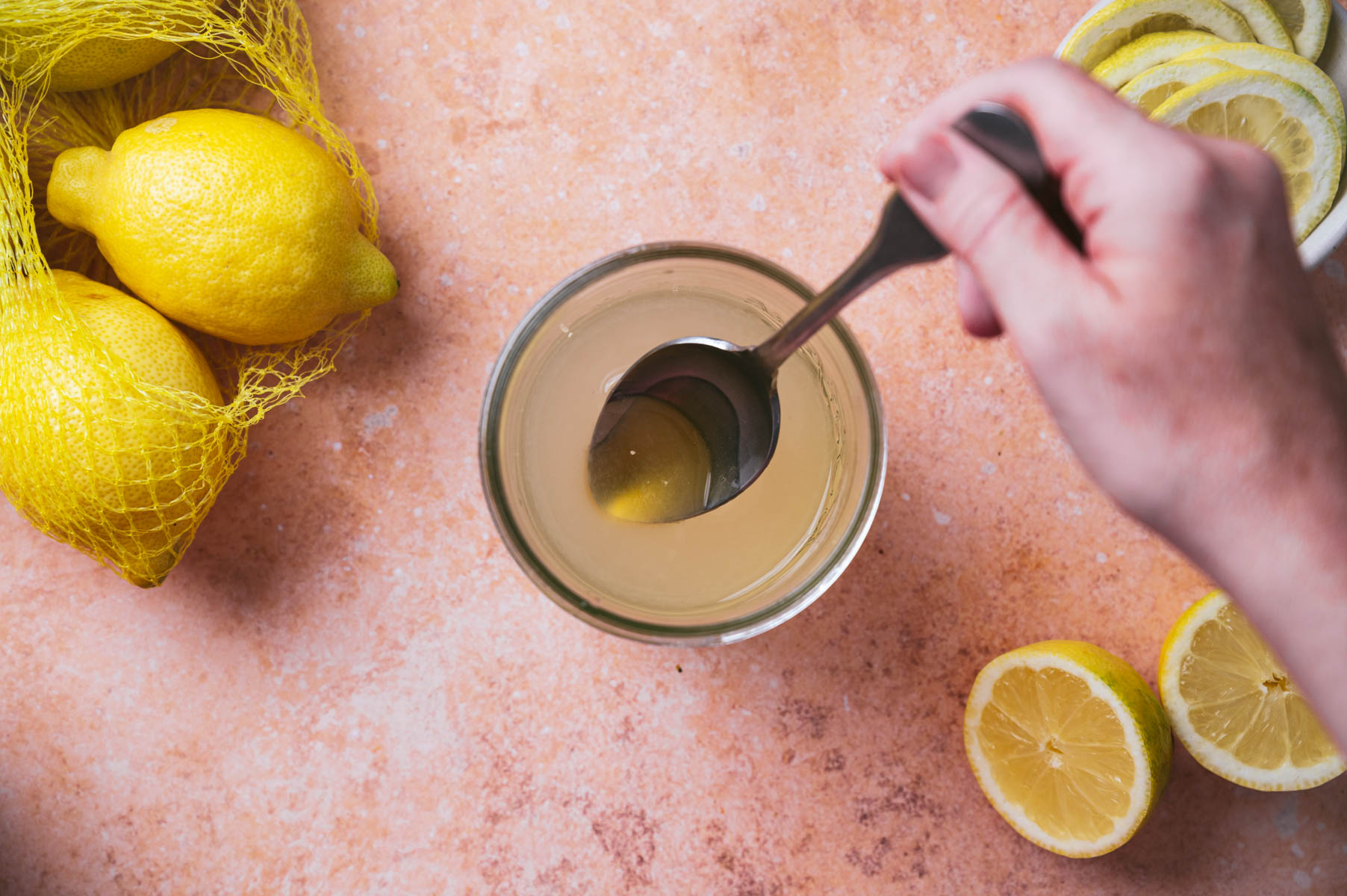 A hand uses a spoon to stir a glass of lemon juice. Lemons in a mesh bag, sliced lemons, and a small bowl with lemon slices are placed on a textured surface.