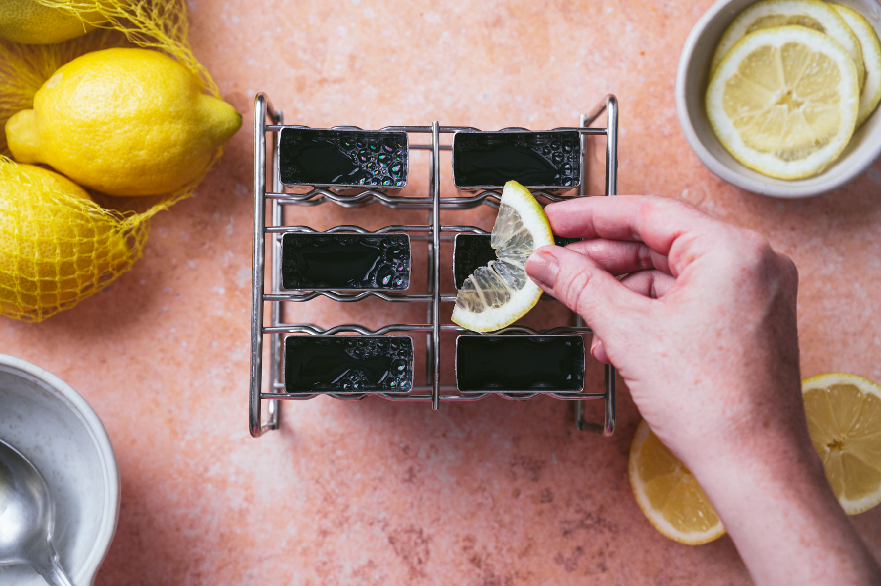 Hand placing a lemon slice on a rack of small black desserts, surrounded by whole lemons, lemon slices, and a bowl on a light pink surface.