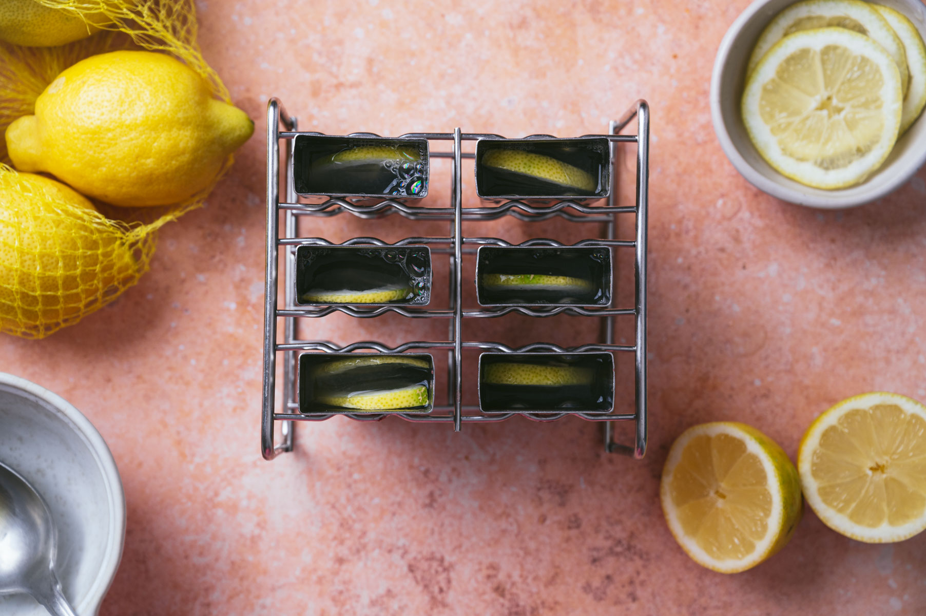 A metal rack holds six square shot glasses filled with a green liquid and garnished with lemon wedges. Lemons and sliced lemon pieces in bowls are placed around the rack.