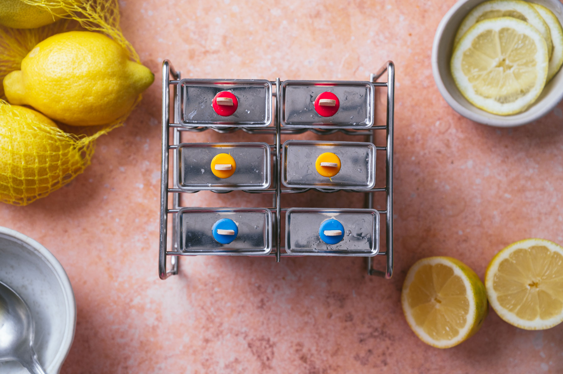 A metal spice rack with six jars, each with a colorful lid, is placed on a pinkish countertop. Surrounding the rack are whole and halved lemons, a bowl with lemon slices, and a partially visible spoon.