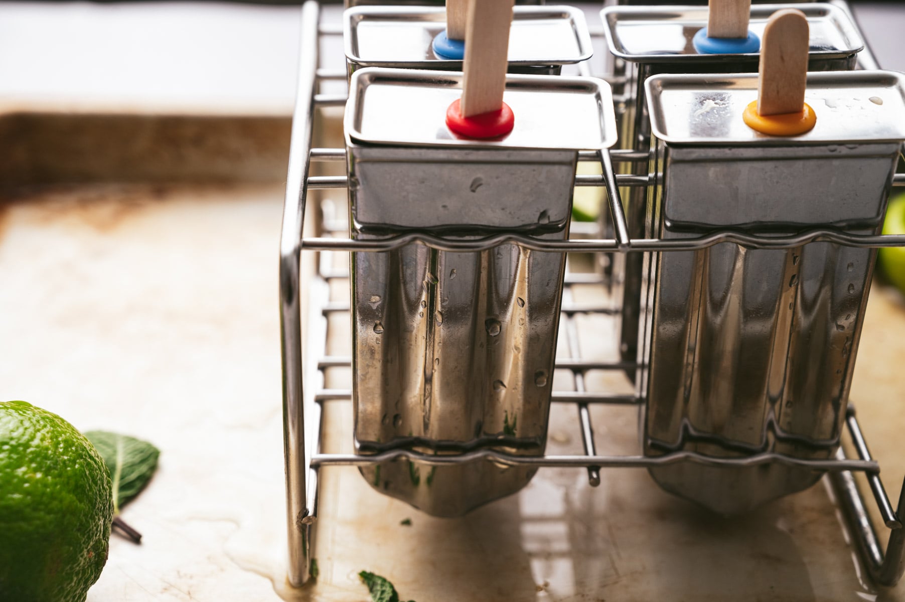 Four metal popsicle molds with wooden sticks are placed on a rack, with a lime and mint leaves nearby on a wooden surface.