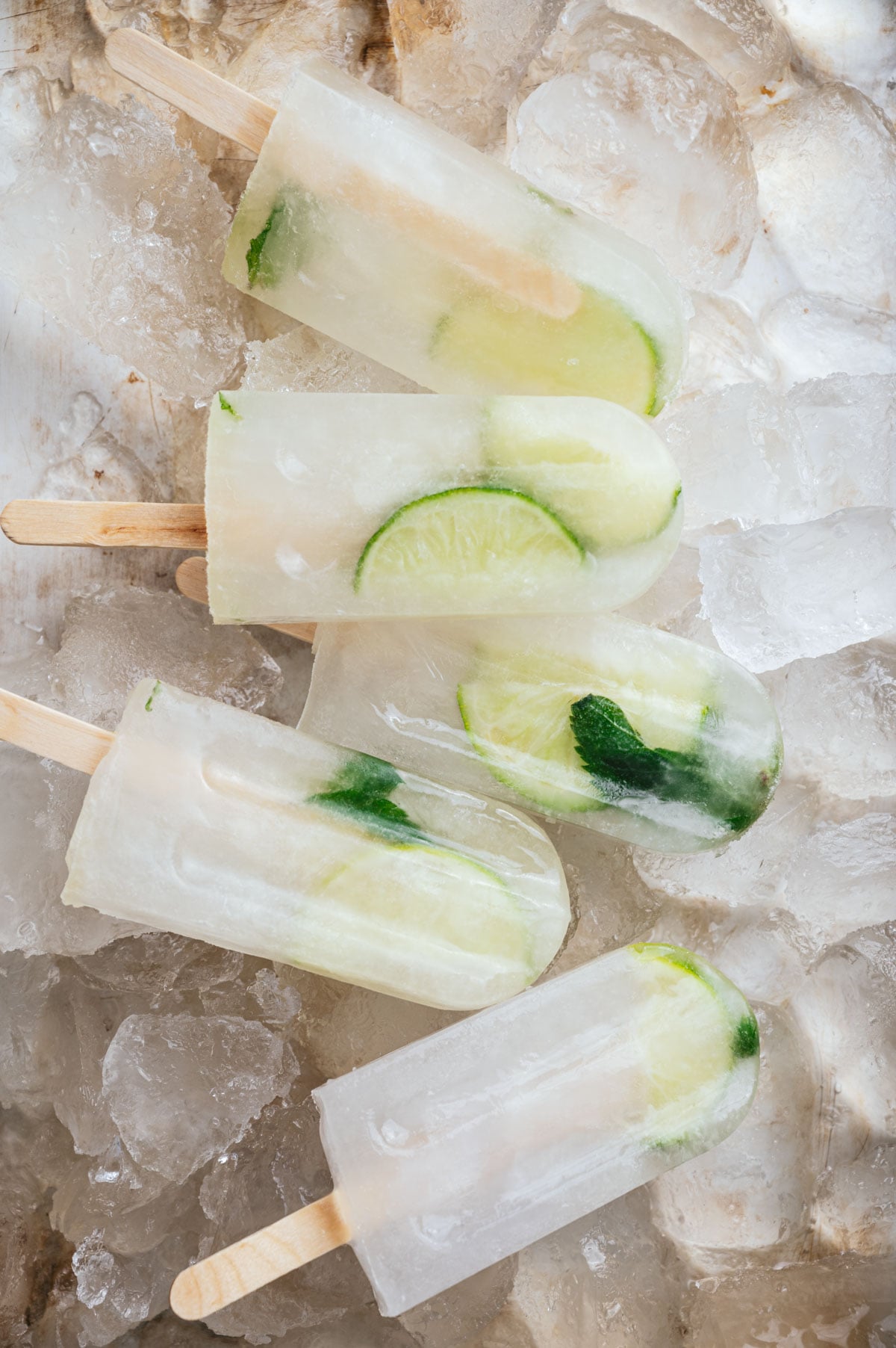 Five lime popsicles with visible lime slices and mint leaves are arranged on a bed of ice cubes.