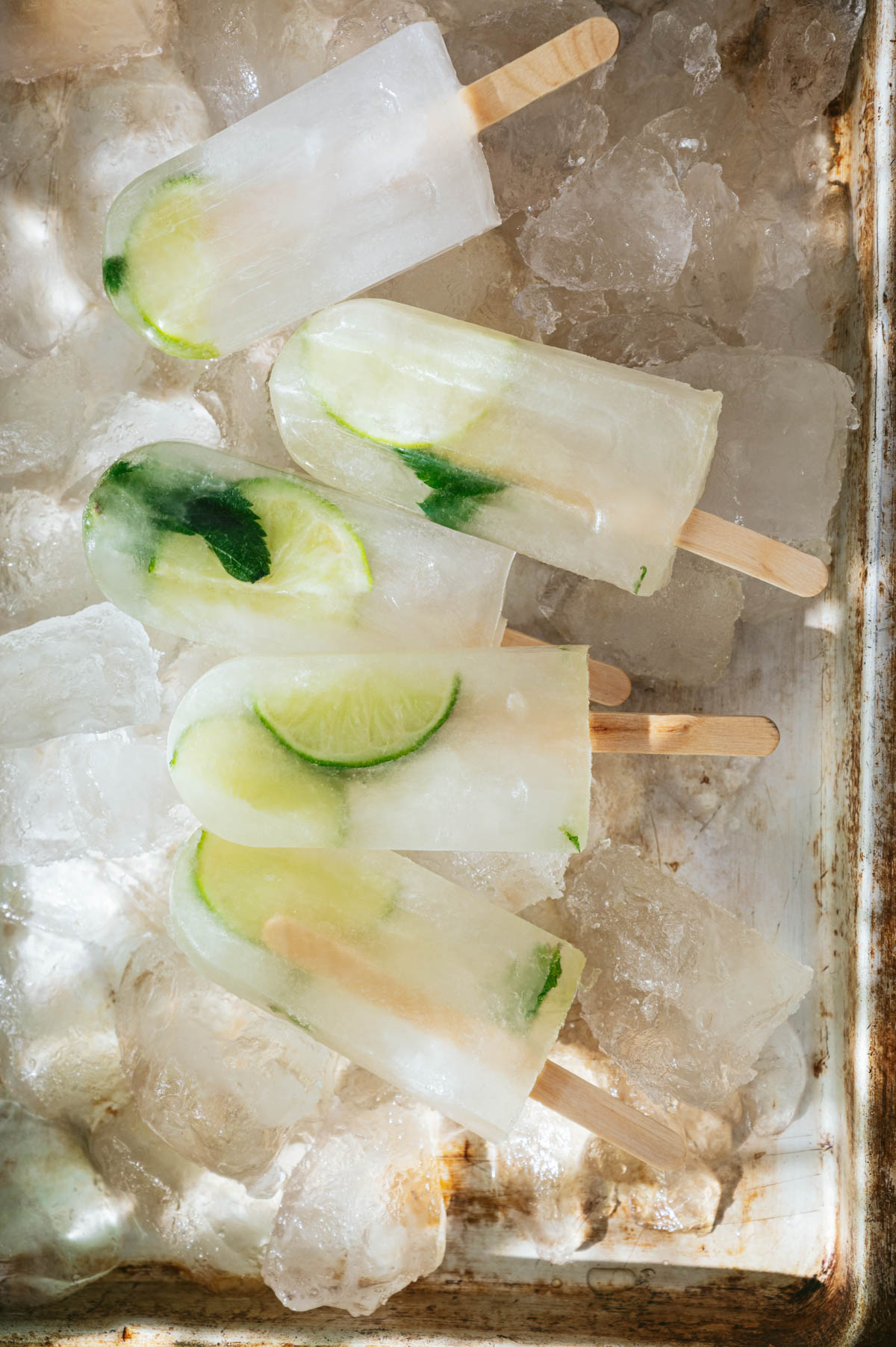 Five lime and mint popsicles are placed on a bed of ice cubes in a metal tray.
