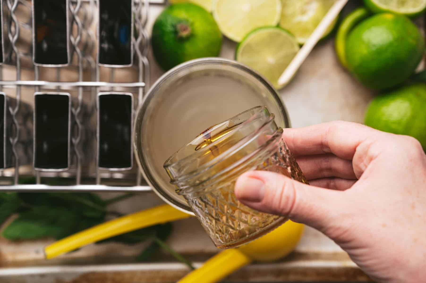 Person adding maple syrup from a small jar to a glass of beverage, surrounded by limes, a yellow citrus juicer, mint leaves, and a utensil holder.