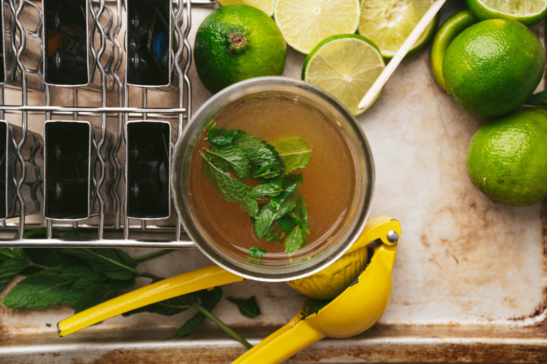 A top-down view of a glass with a brown liquid and mint leaves, surrounded by lime halves, a yellow citrus squeezer, and a rack holding multiple shot glasses.