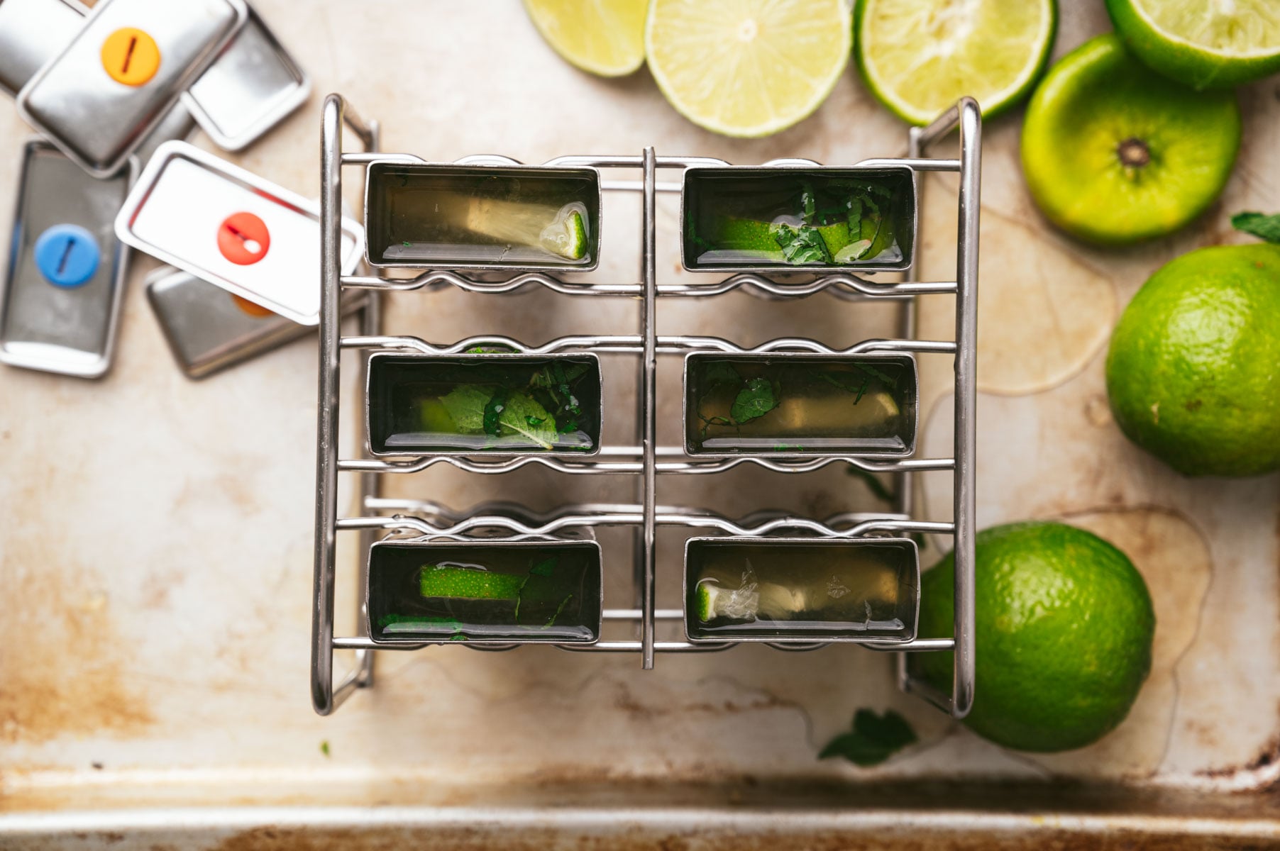Metal ice pop molds filled with lime and mint, arranged in a metal holder. Whole and sliced limes are scattered around the molds on a countertop.