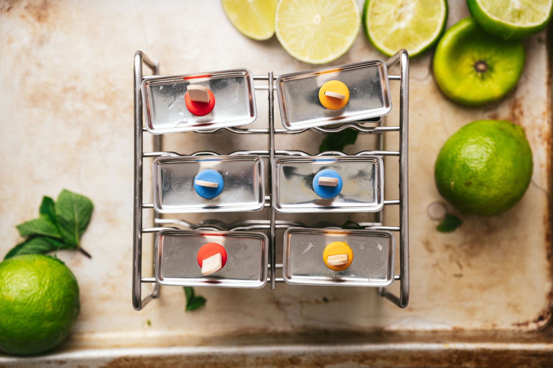 A glass rack holds six bottles sealed with colorful caps. Surrounding the rack are sliced and whole limes on a light-colored surface with mint leaves.