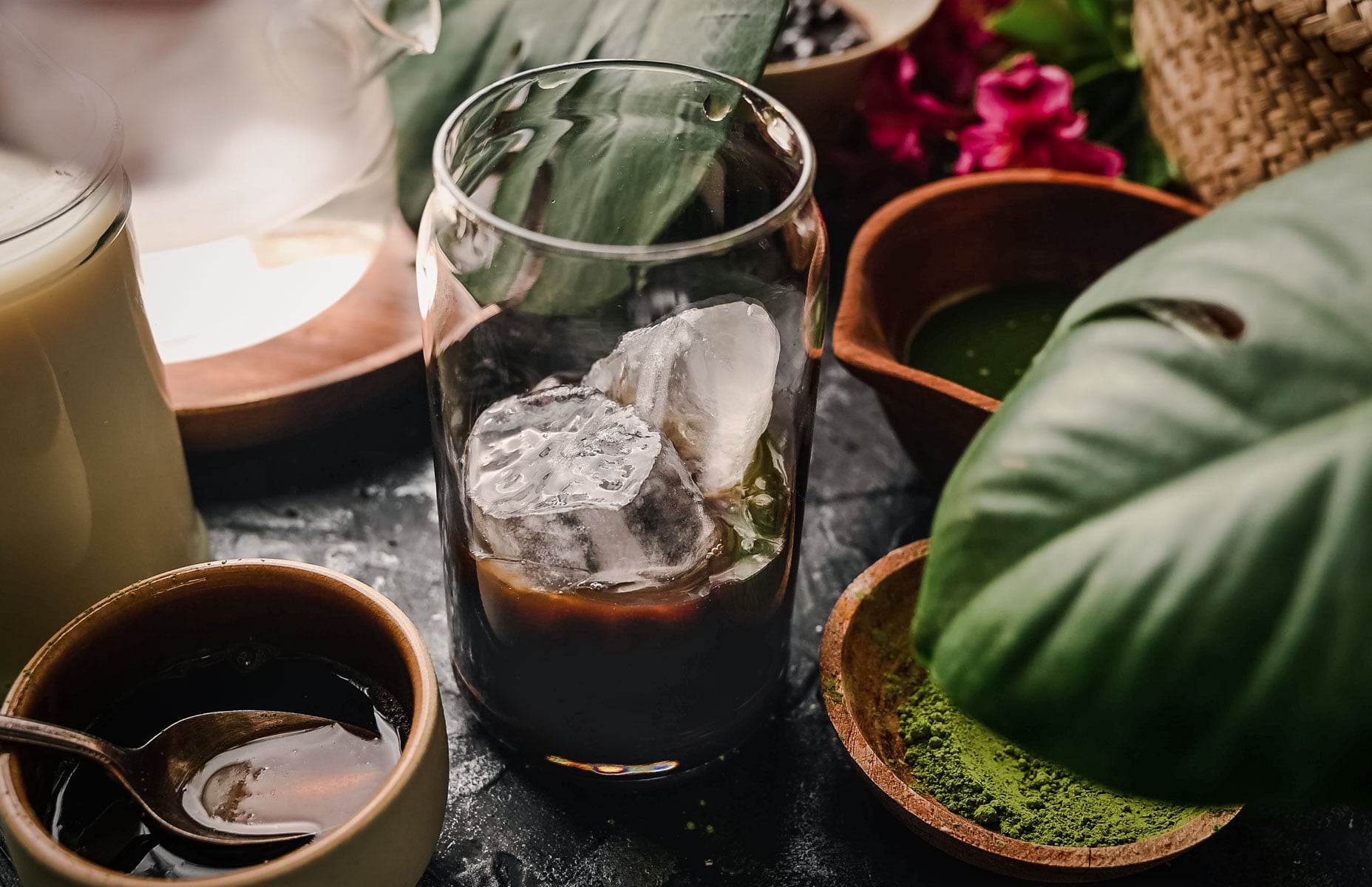 A glass of iced coffee is surrounded by bowls containing ingredients like matcha powder, a spoon in a dark liquid, and greenery, set on a dark surface.