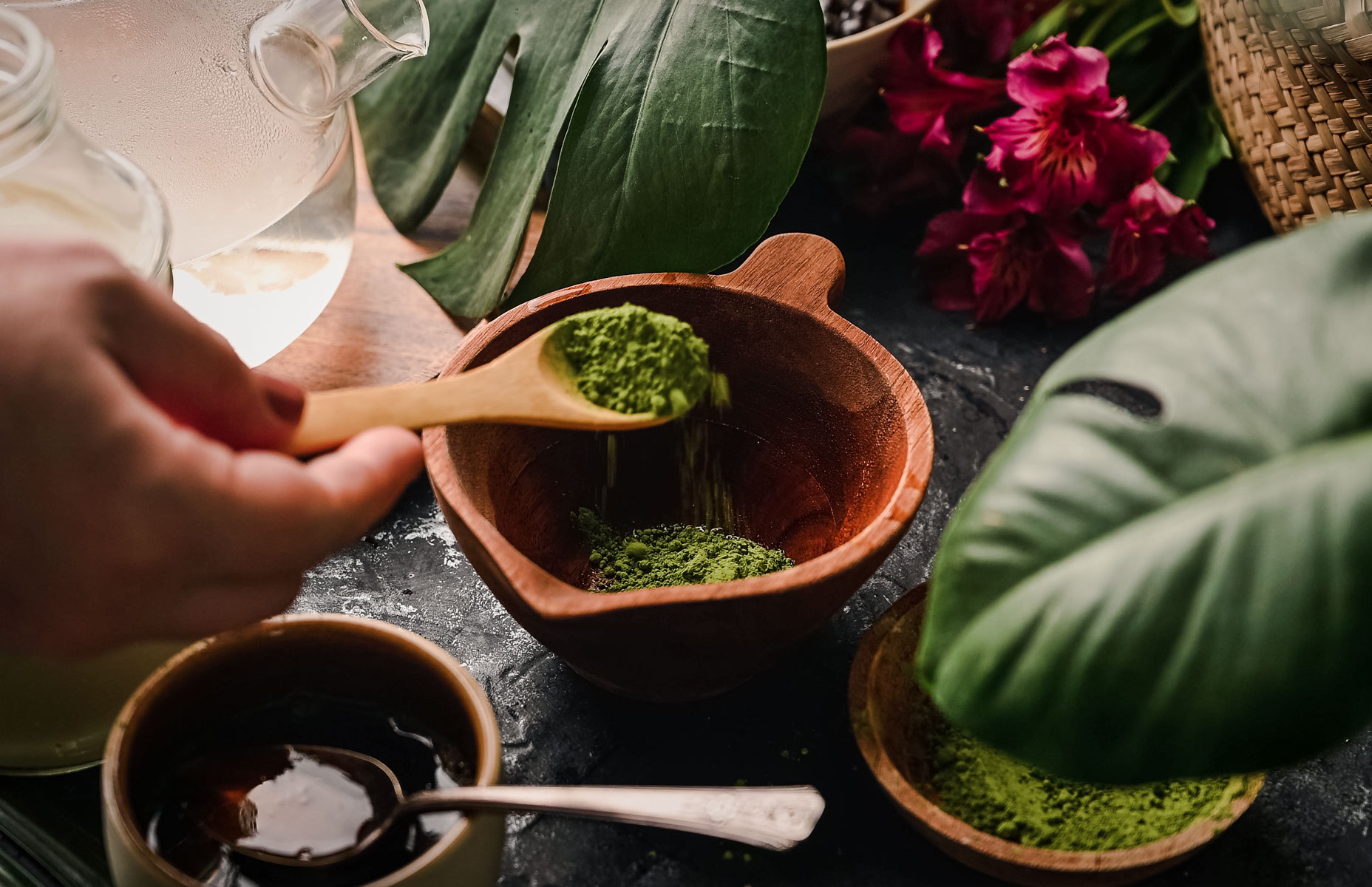 A hand holding a wooden spoon sprinkles green tea matcha powder into a wooden bowl. Nearby are a cup of dark beverage, a clear teapot, and green leafy plants. Bright red flowers are in the background.