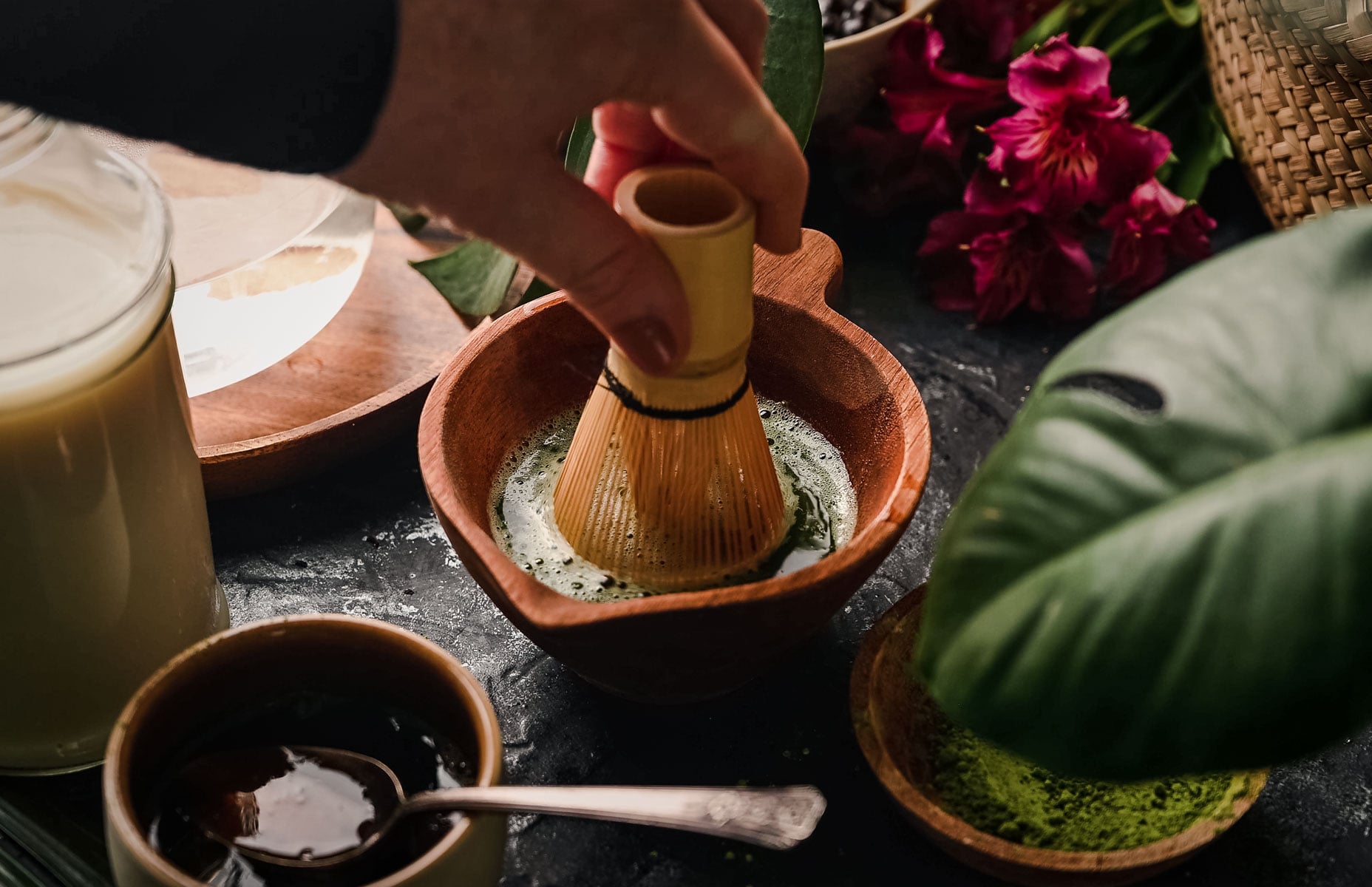 A hand uses a bamboo whisk to stir matcha in a wooden bowl, with tea accessories and green leaves nearby.