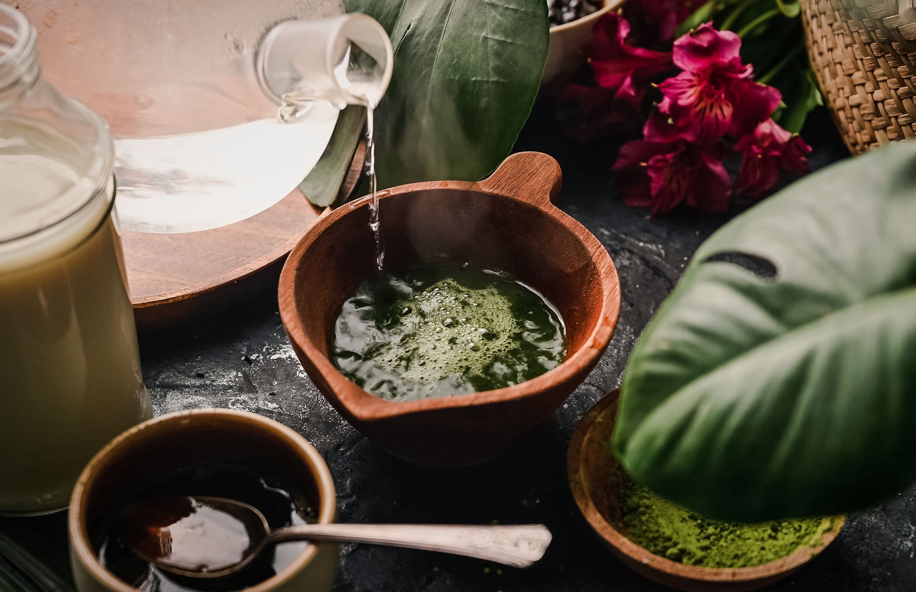 Water is being poured into a wooden bowl containing green matcha powder, surrounded by leaves, flowers, and a jar with liquid on a dark surface.