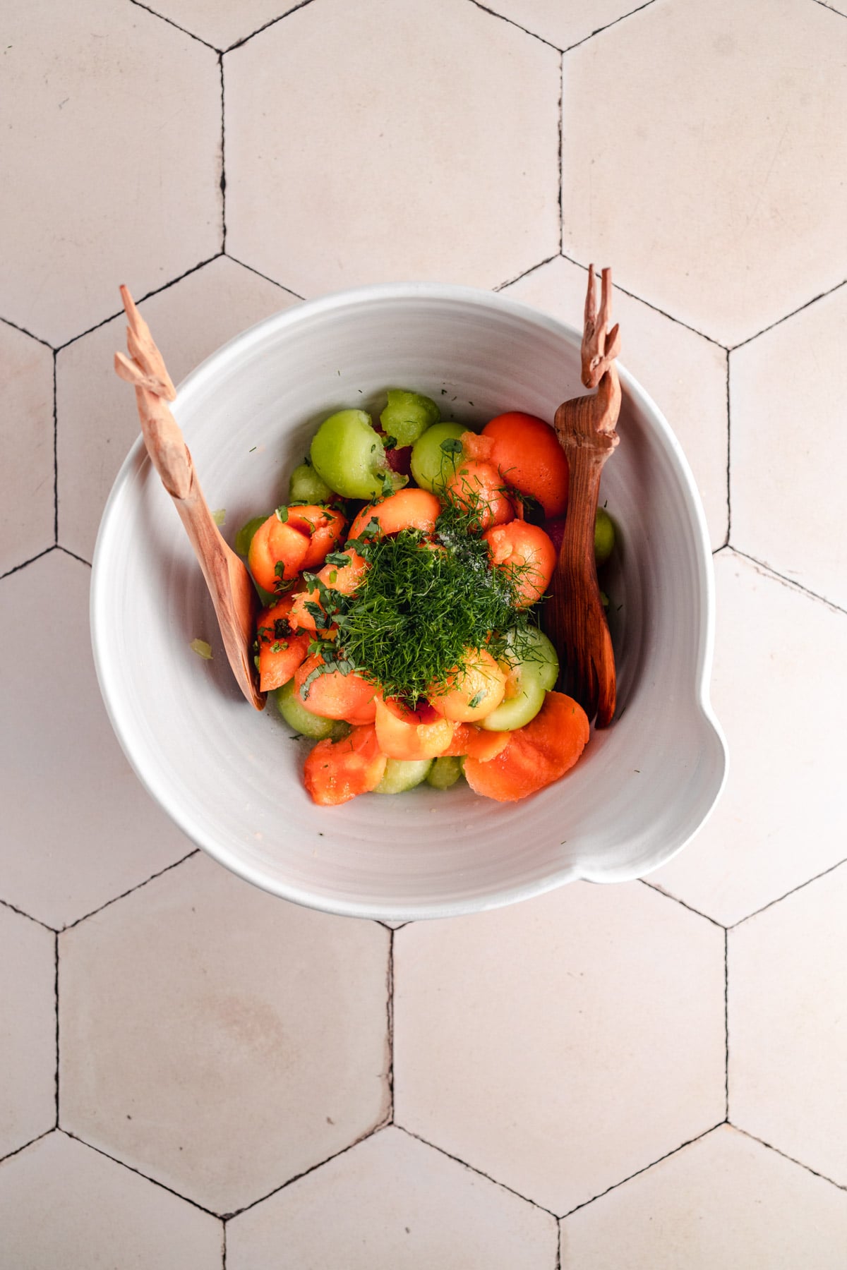 A white bowl containing a melon and carrot salad garnished with herbs and accompanied by two wooden utensils, placed on a hexagon-patterned tile surface.