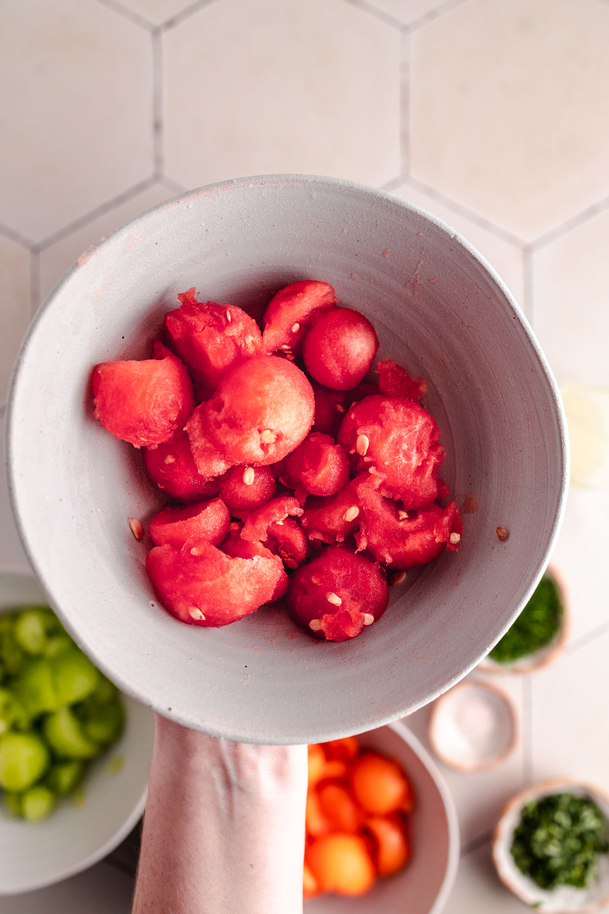 A hand holding a bowl filled with scooped watermelon pieces. Additional bowls with green and orange fruits are visible in the background.