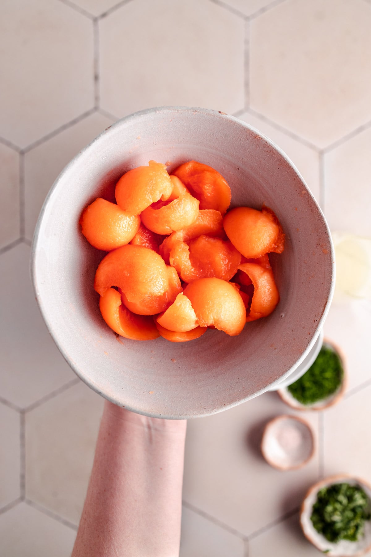 A bowl containing orange melon balls, being held by a hand over a tiled surface background. Small dishes with herbs and other ingredients are seen below the bowl.