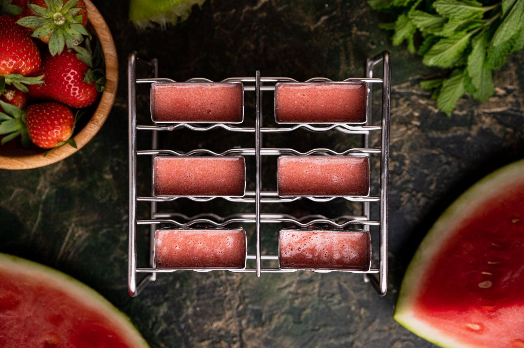 Six strawberry watermelon ice pops in a metal mold, surrounded by fresh strawberries, mint leaves, and watermelon slices.
