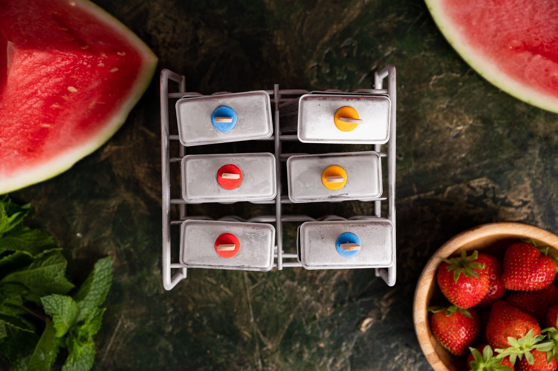 A top view of an ice pop mold tray with six popsicles, surrounded by watermelon slices, strawberries in a bowl, and mint leaves on a dark marble surface.