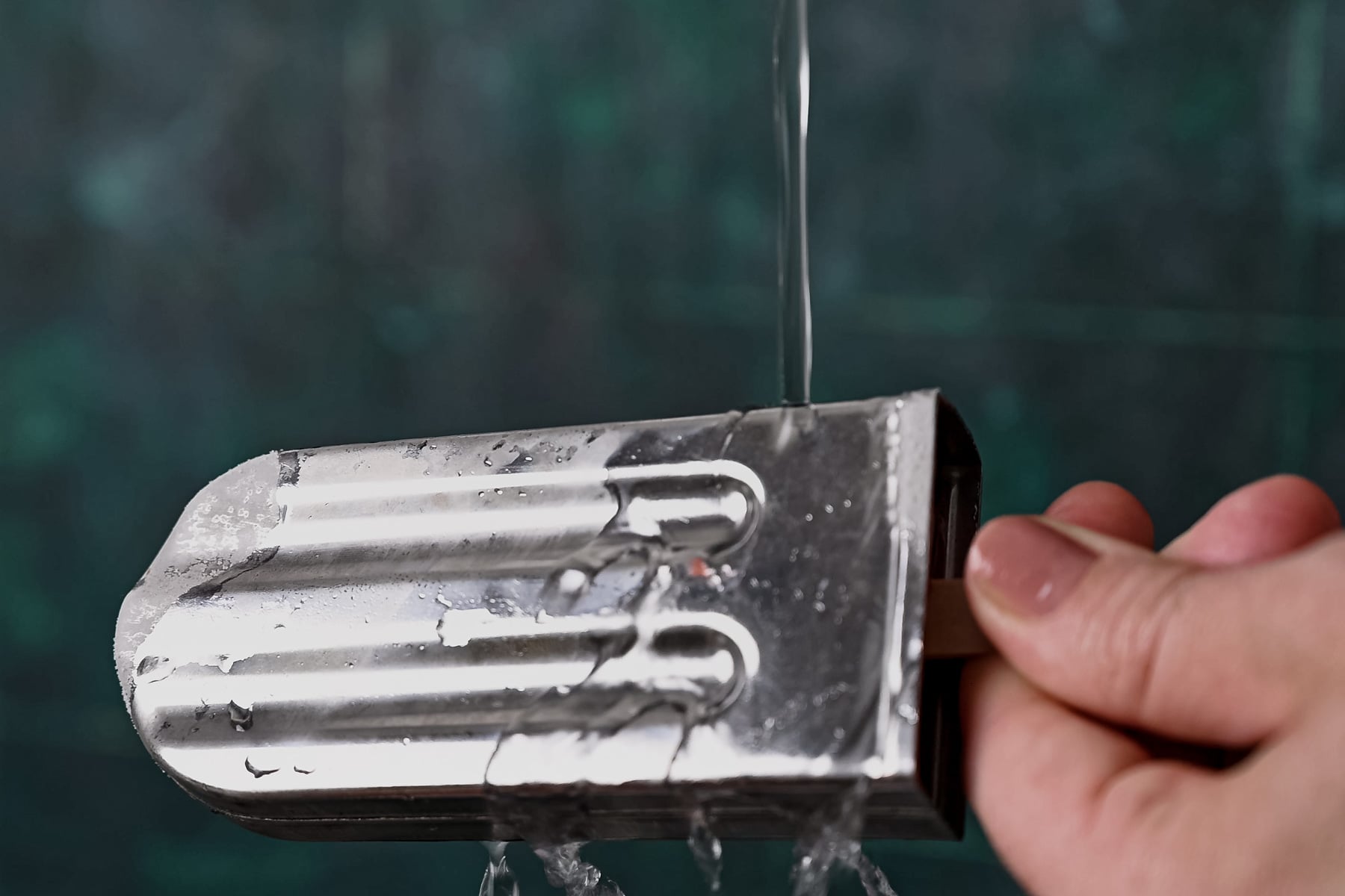 A hand is holding a silver metal popsicle mold under running water, with the background blurred.