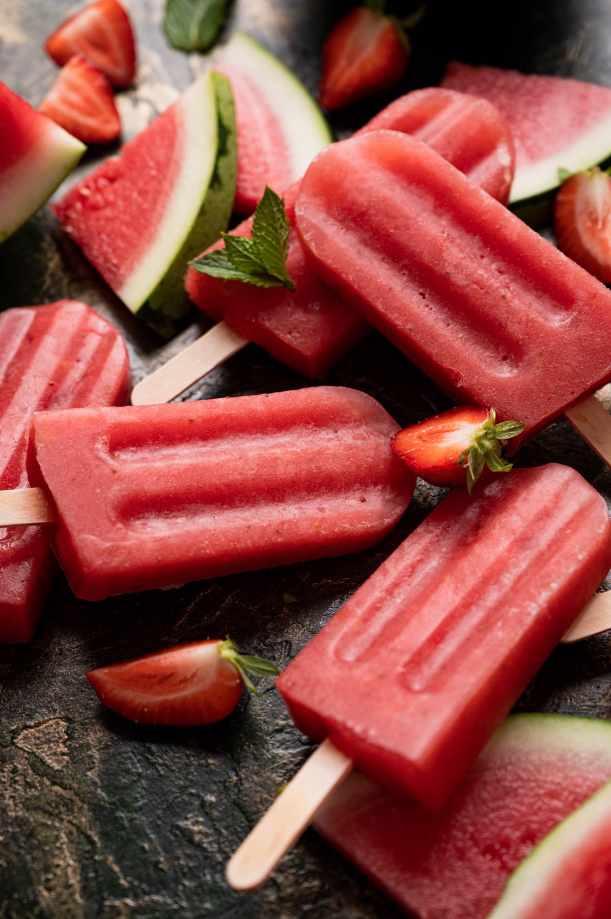 A variety of watermelon and strawberry popsicles, garnished with fresh strawberry slices and mint leaves, are placed on a dark surface.